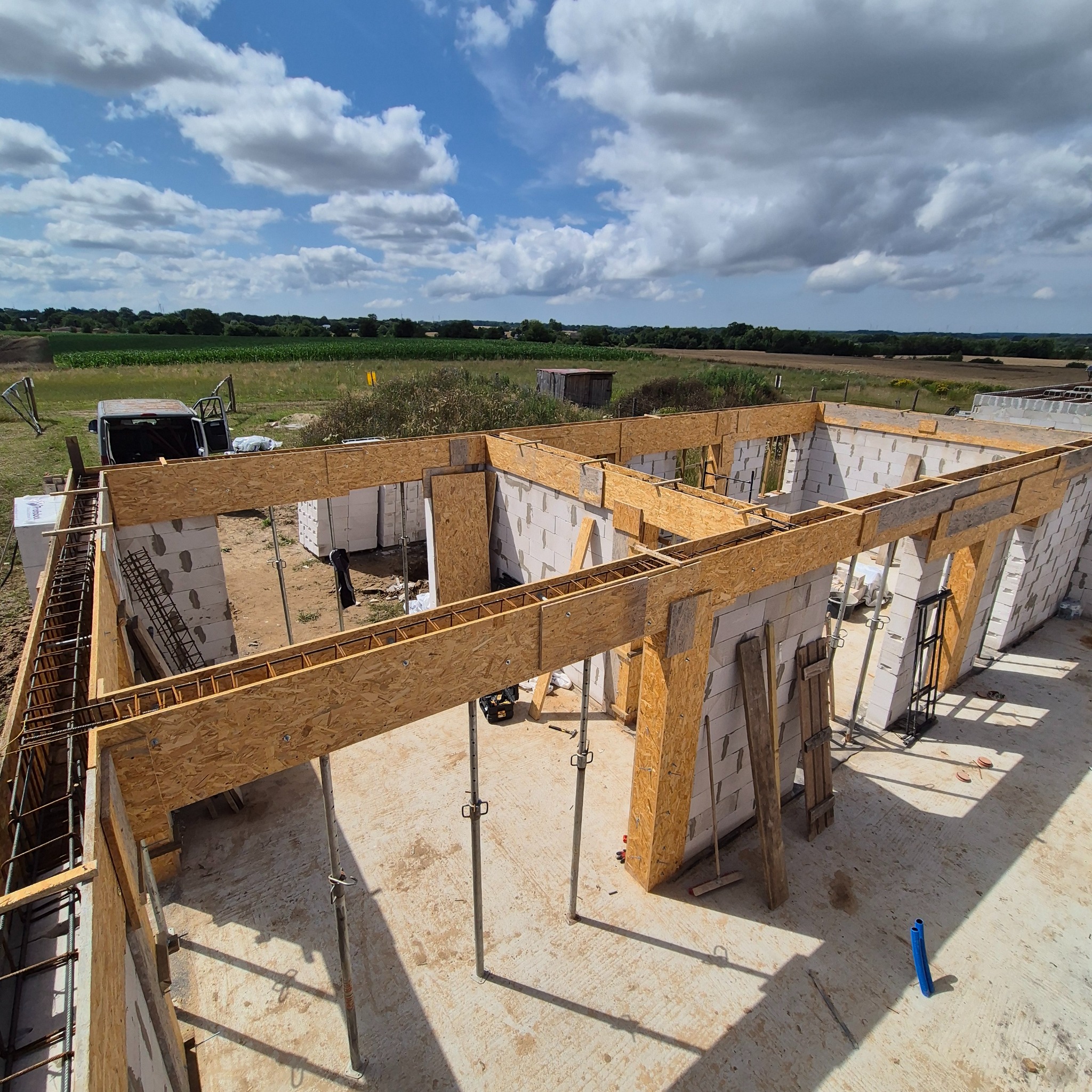 Wznoszenie ścian domu z betonu komórkowego. Widoczna konstrukcja szalunkowa z drewna i zbrojenie. Na zewnątrz krajobraz z polami i częściowo zachmurzone niebo.