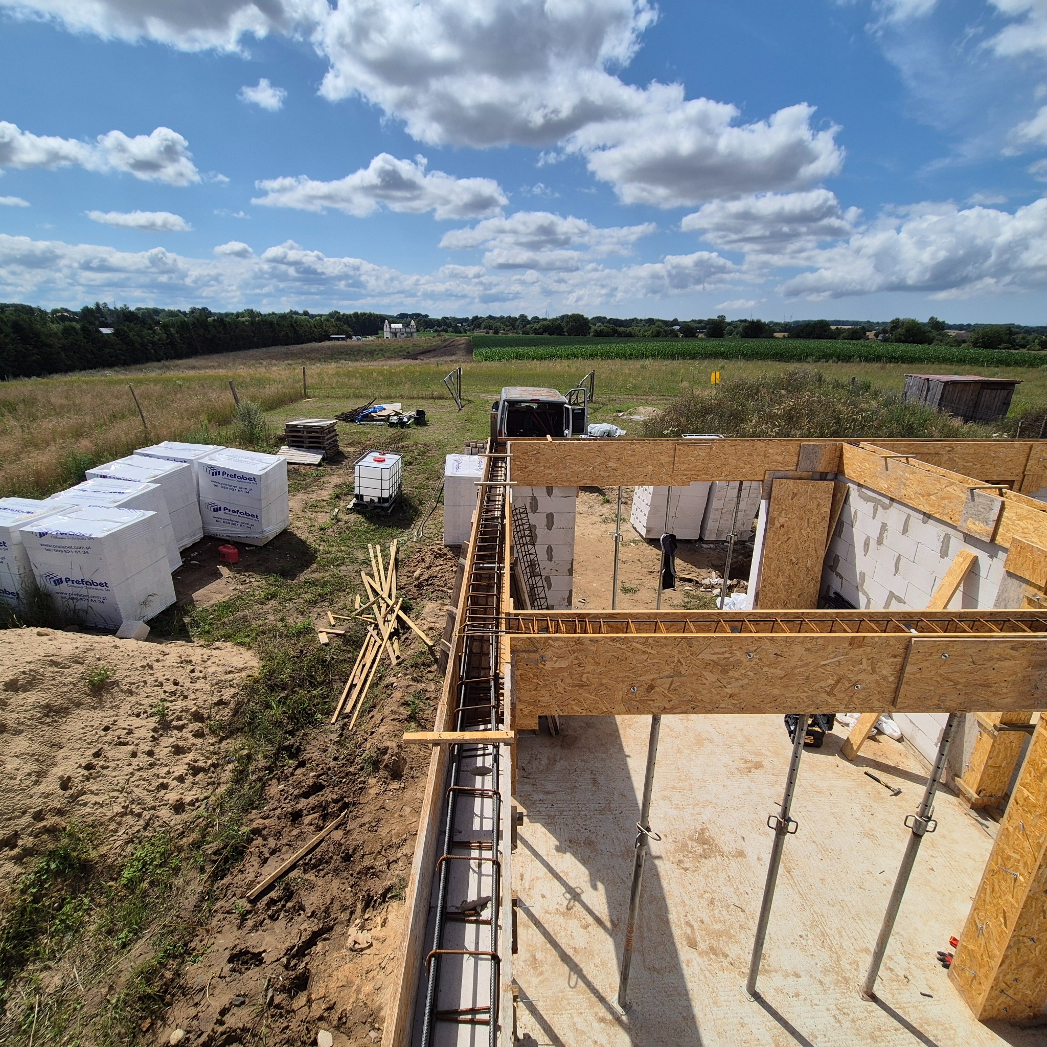 Budowa domu z bloczków betonu komórkowego: fundamenty zbrojone, ściany w trakcie wznoszenia, materiały budowlane na placu budowy, błękitne niebo z chmurami.