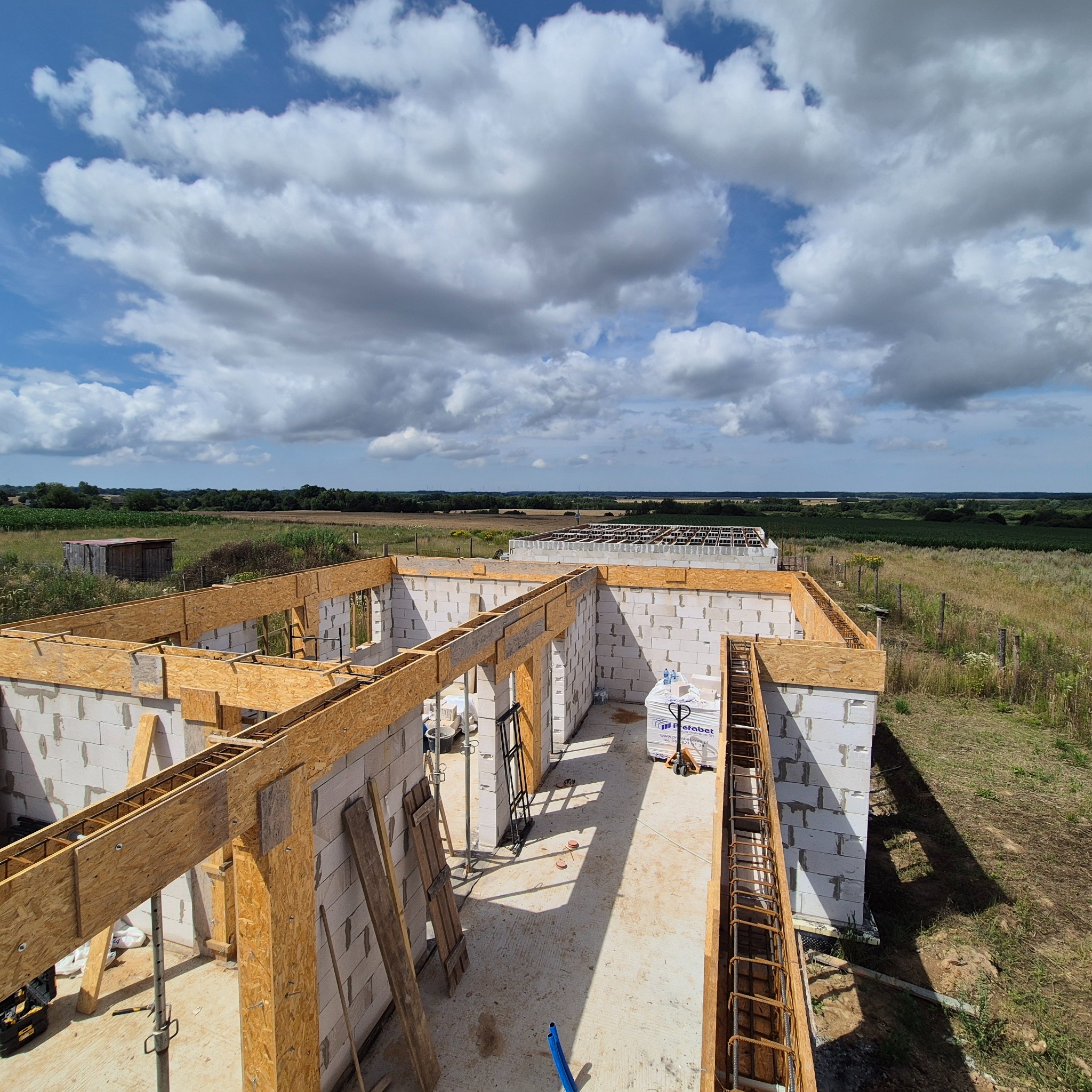 Budowa domu z betonu komórkowego w toku. Widok z góry na ściany, szalunki i zbrojenia. W tle krajobraz i niebo z chmurami. Etap realizacji.