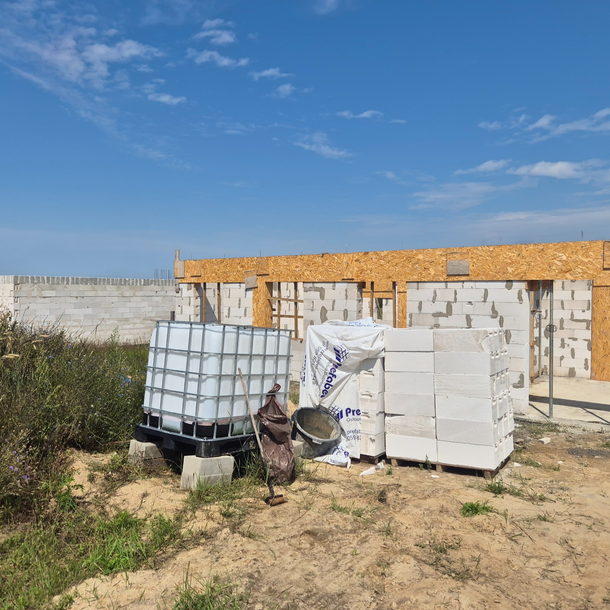 Budowa domu z betonu komórkowego w Manowie. Widoczne ściany w trakcie murowania, zbiornik na wodę i materiały budowlane.
