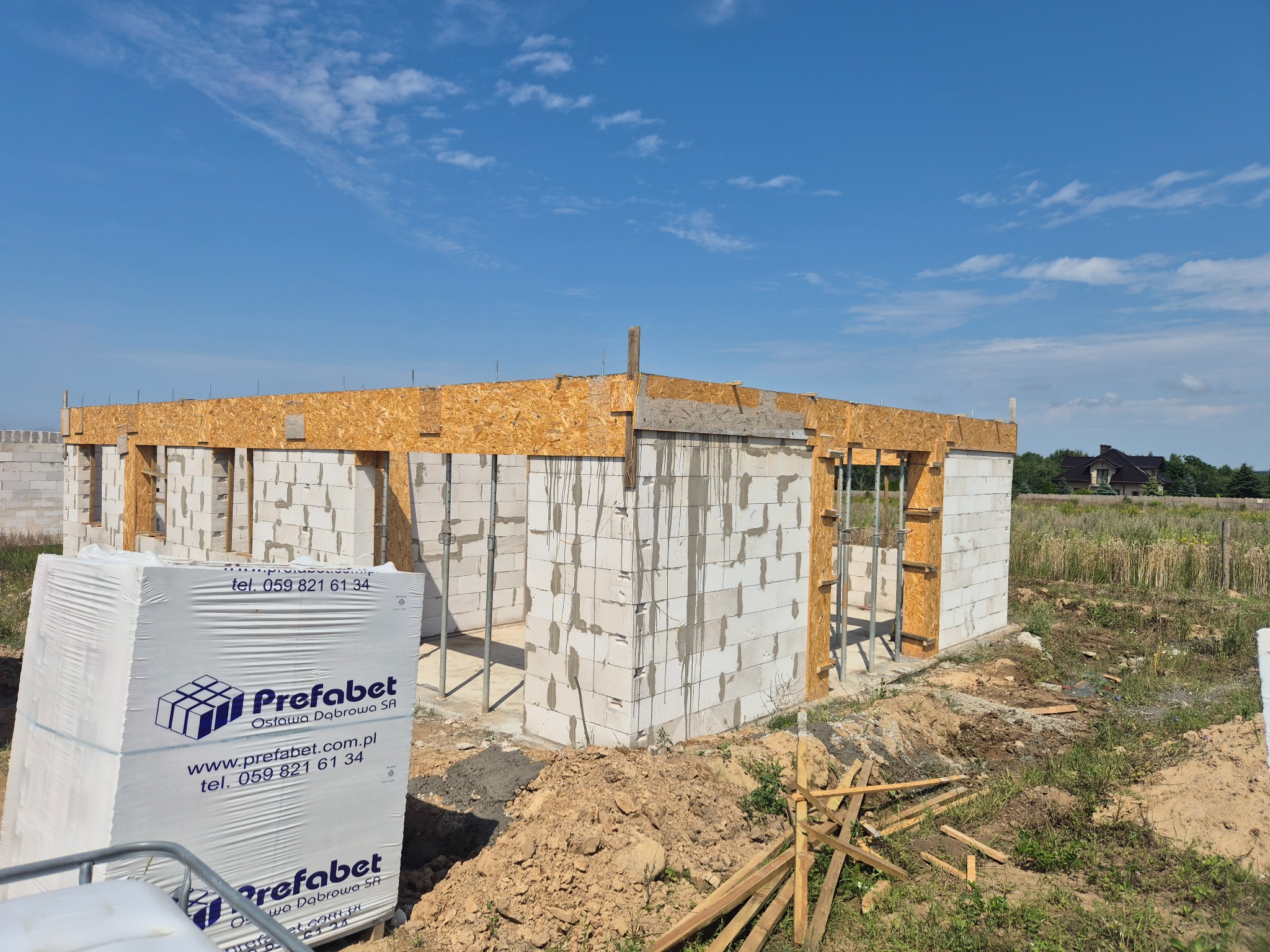 Budowa domu z bloczków Prefabet: wzniesione ściany parteru z drewnianą konstrukcją szalunkową. Na pierwszym planie paleta bloczków. Błękitne niebo w tle.