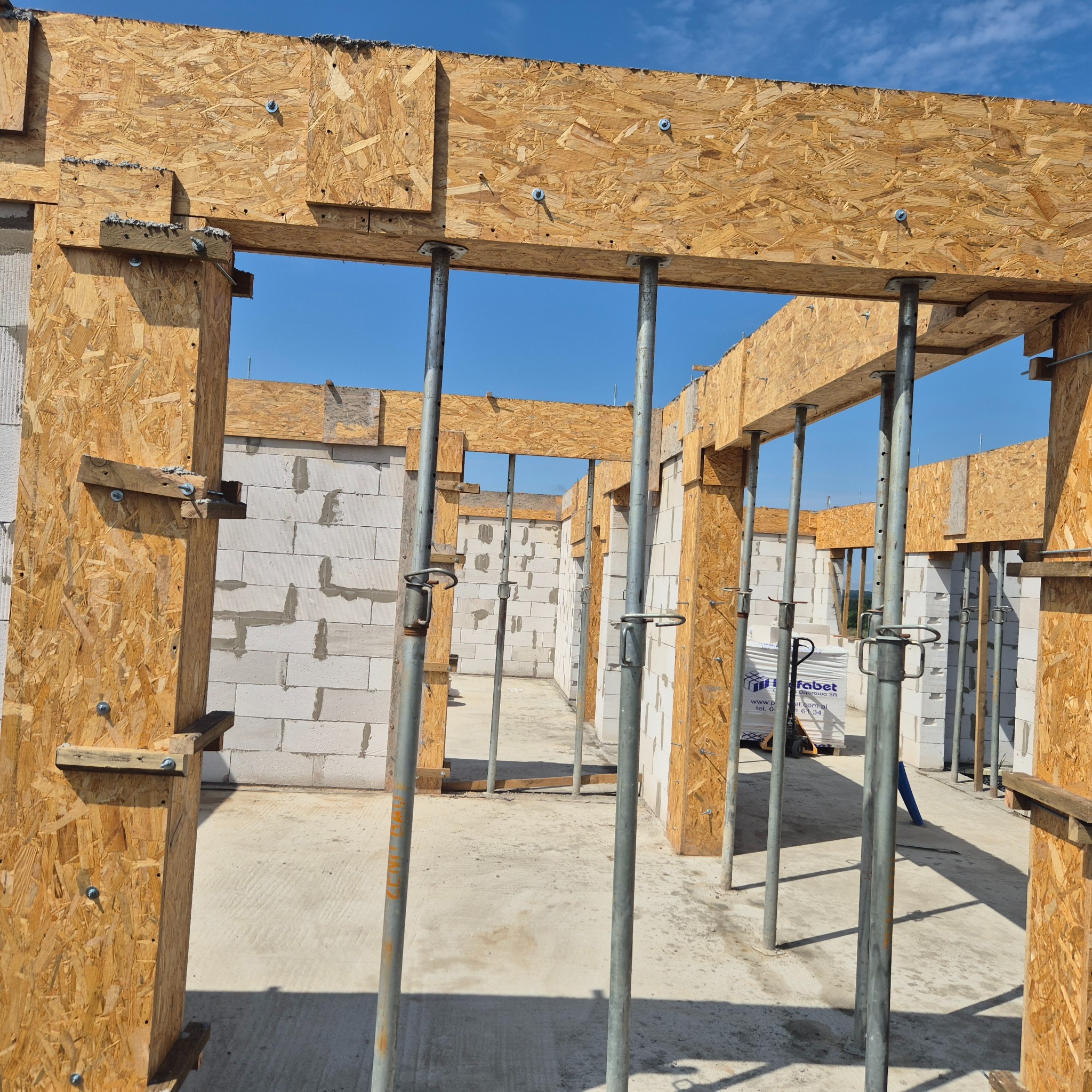 Wznoszenie ścian z bloczków betonu komórkowego, wspartych szalunkiem z płyty OSB i metalowymi stemplami. Widoczne palety z bloczkami w głębi budynku.