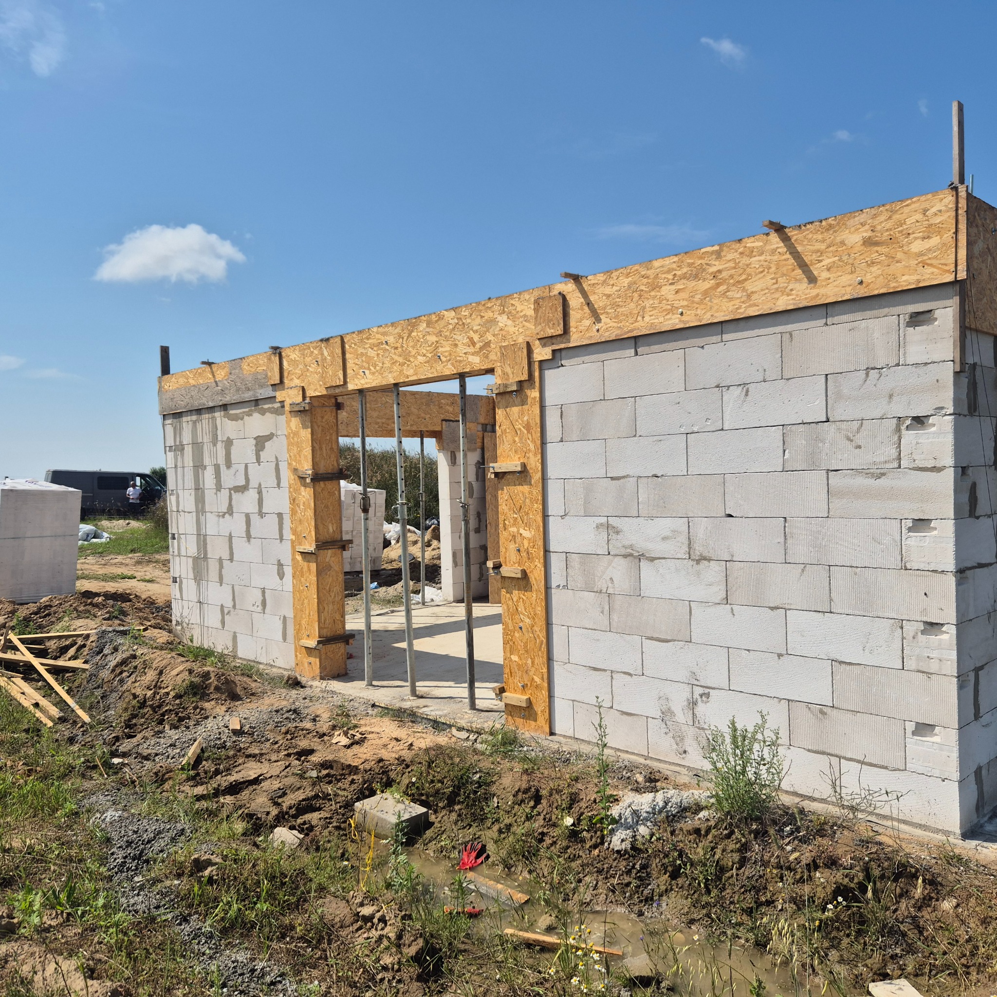 Wznoszenie ścian domu z betonu komórkowego. Widoczne drewniane szalunki okienne i drzwiowe. Na zewnątrz budynku leżą bloczki i widać samochód dostawczy.