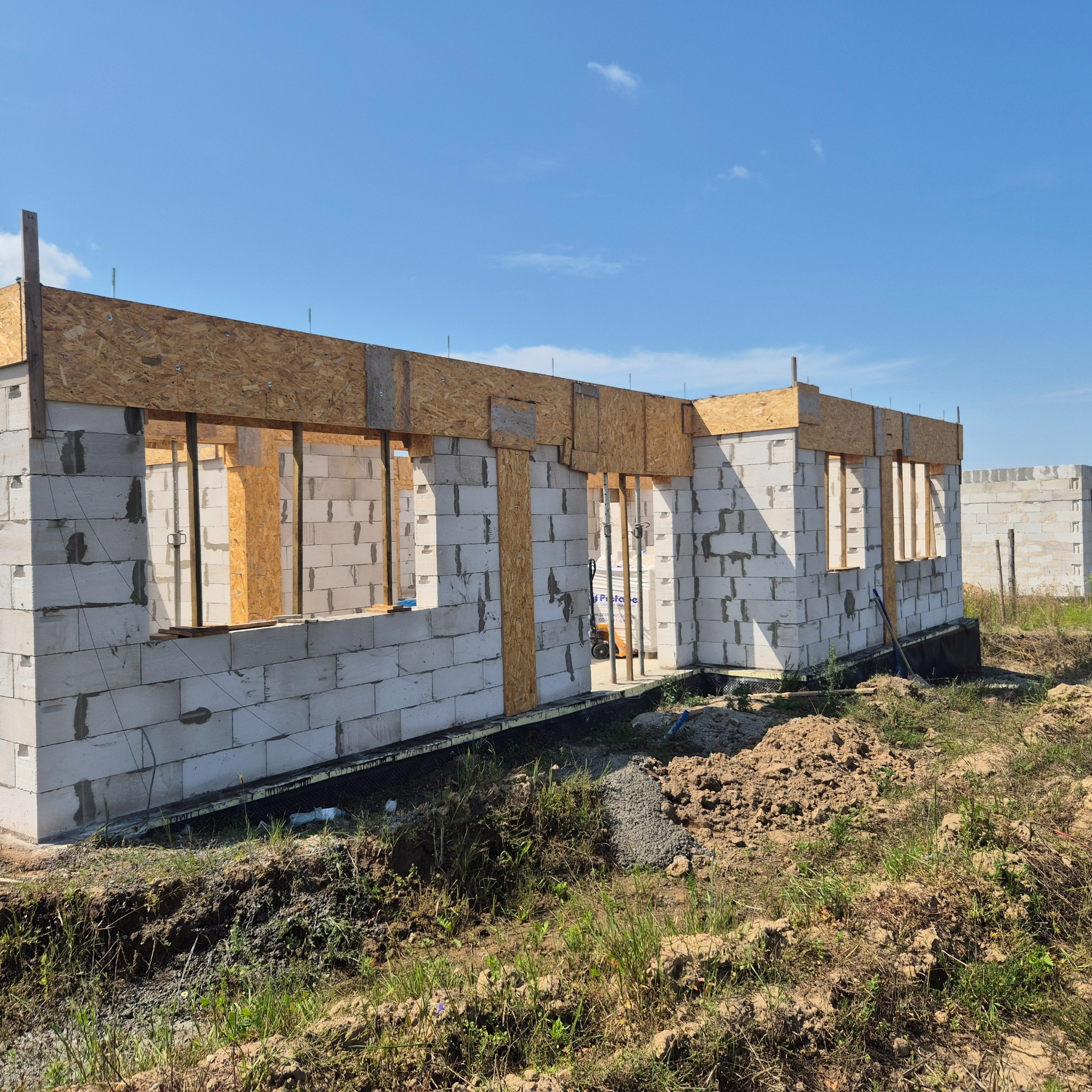 Budowa domu w trakcie: ściany z betonu komórkowego, drewniane nadproża okienne, widoczny fundament i wykop pod instalacje. Błękitne niebo w tle.