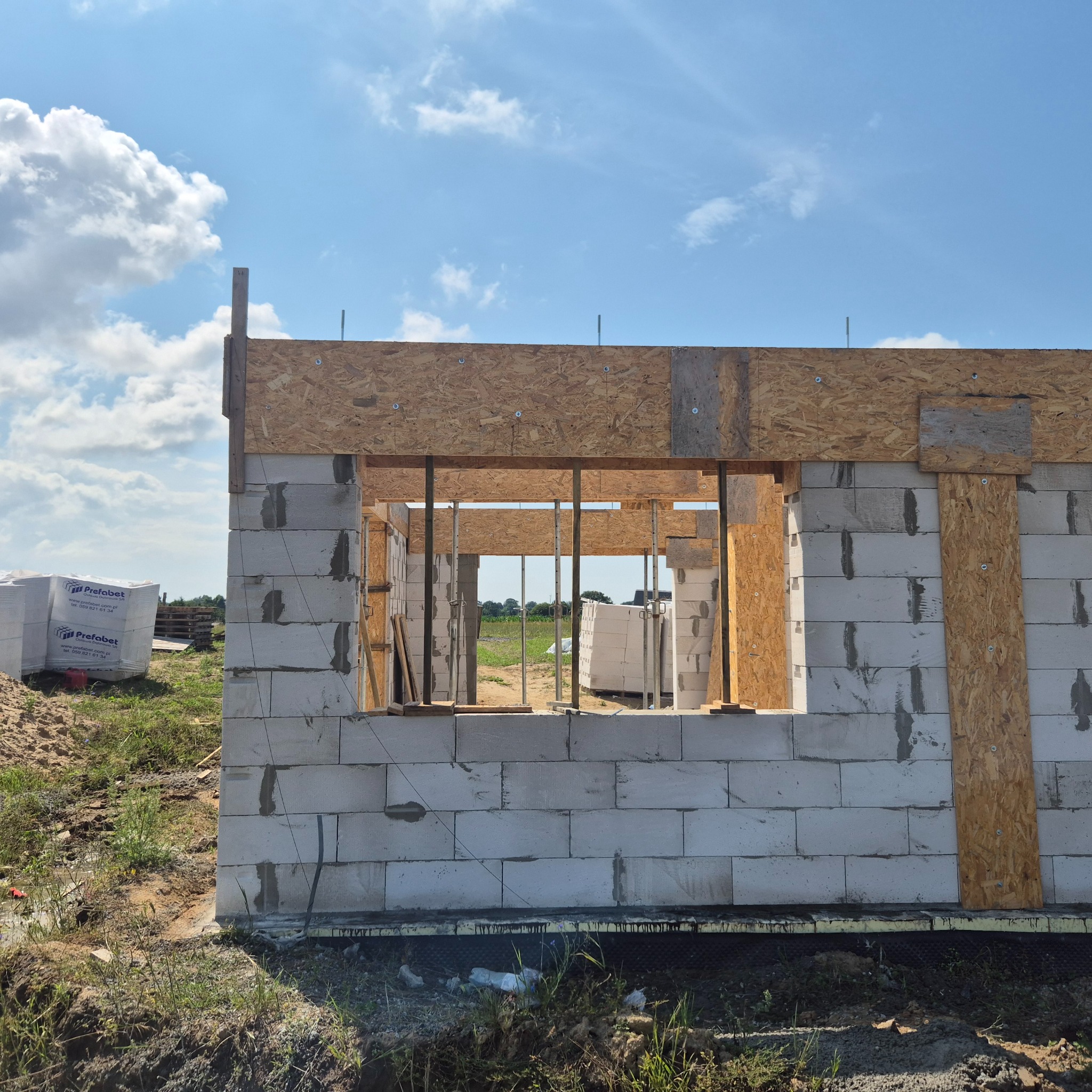 Wznoszenie ścian domu z betonu komórkowego, widoczne szalunki okienne z płyt OSB i zapas materiału budowlanego na placu. Częściowo zrealizowany etap budowy.