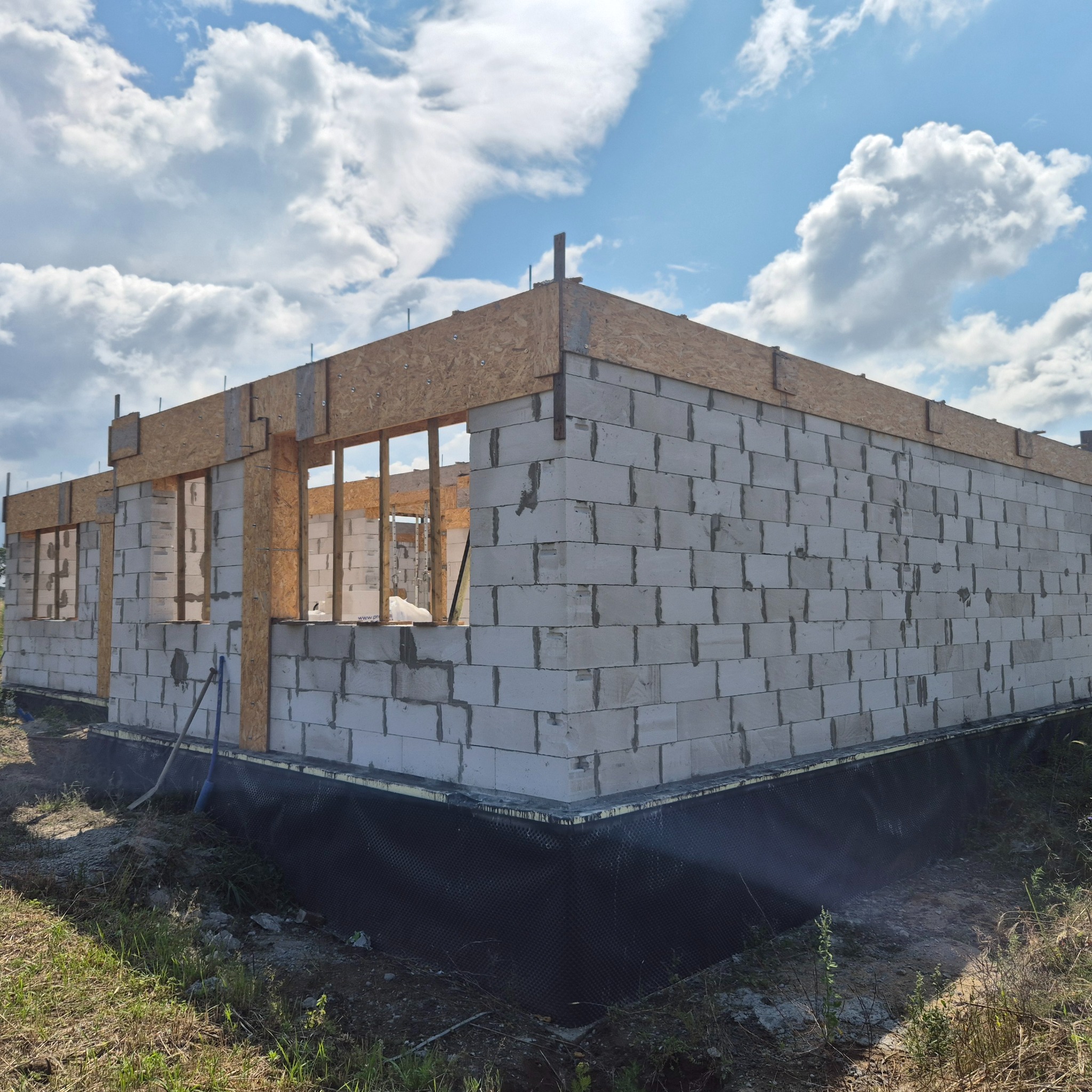 Budowa domu w toku: ściany z betonu komórkowego, drewniane nadproża okien, widoczna izolacja fundamentów. Błękitne niebo z chmurami w tle.