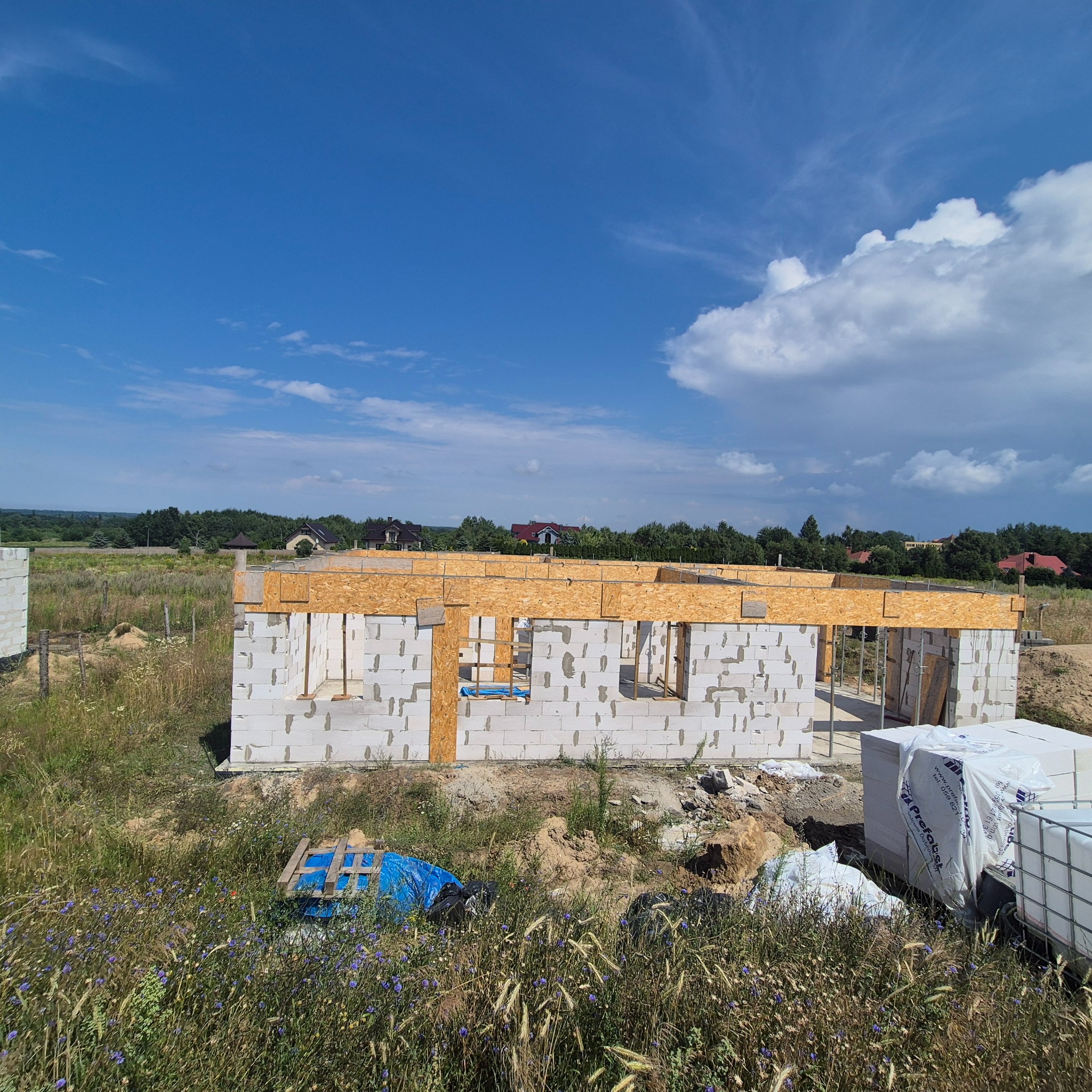 Wznoszenie ścian parterowego domu z betonu komórkowego. Częściowo ukończona konstrukcja z drewnianymi nadprożami na tle błękitnego nieba i zieleni.