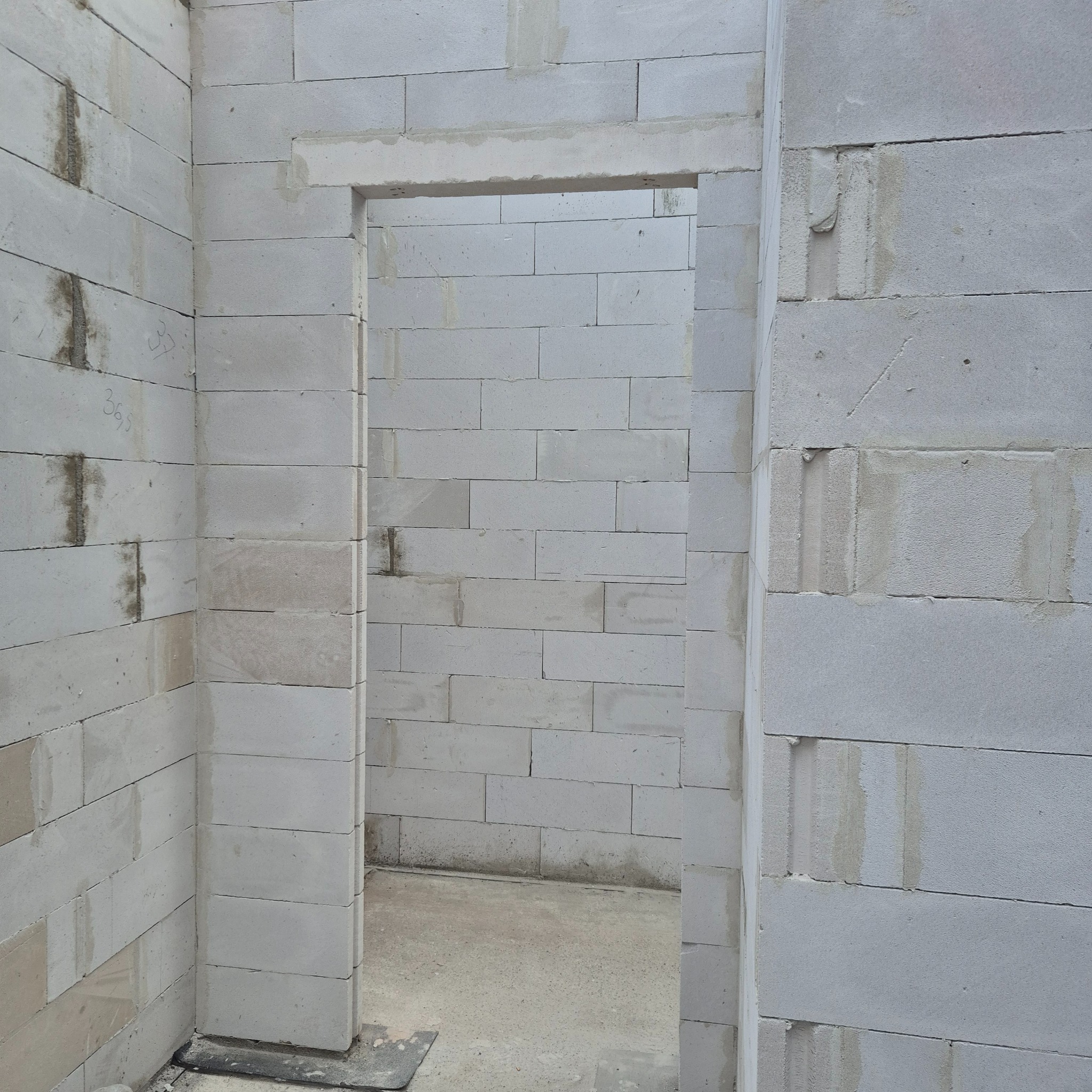 Wnętrze budynku w trakcie budowy: ściany z białych bloczków, widoczne otwory drzwiowe. Na ścianie delikatne ślady zaprawy. Surowy stan, etap murowania.