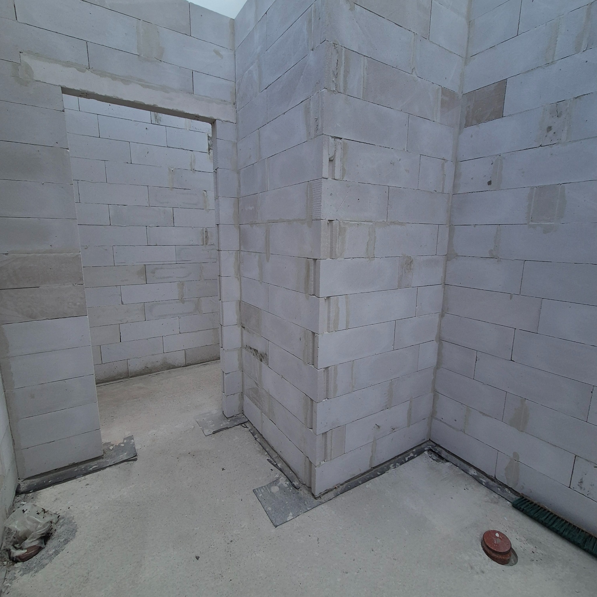 Wnętrze w trakcie budowy: ściany z białych bloczków, widoczne otwory drzwiowe, surowy beton na podłodze. Etap murowania ścian w nowym domu.