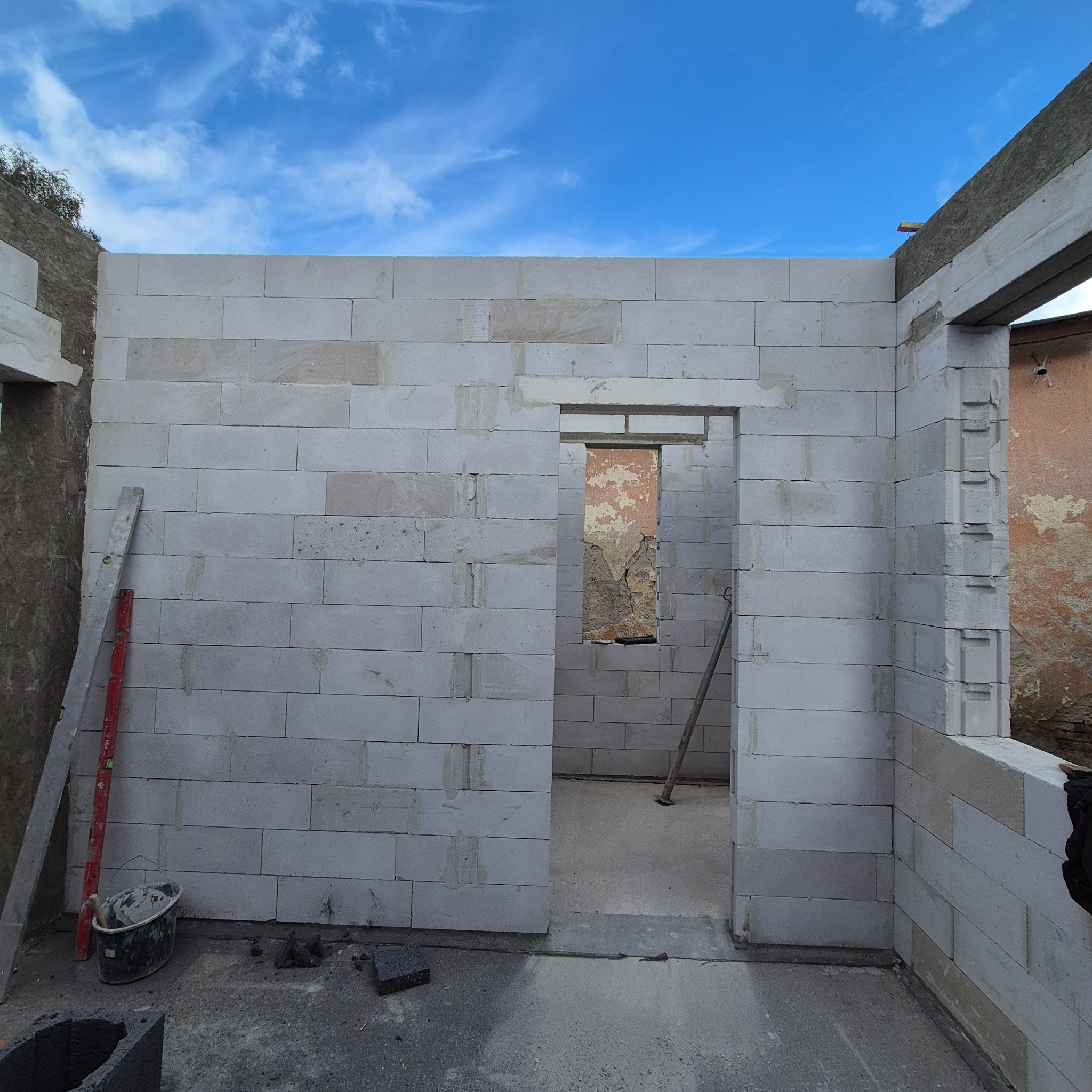 Ściana z białych bloczków, widoczny otwór drzwiowy i okienny. W tle błękitne niebo. Na pierwszym planie poziomica i wiadro. Etap budowy domu z widocznymi narzędziami.