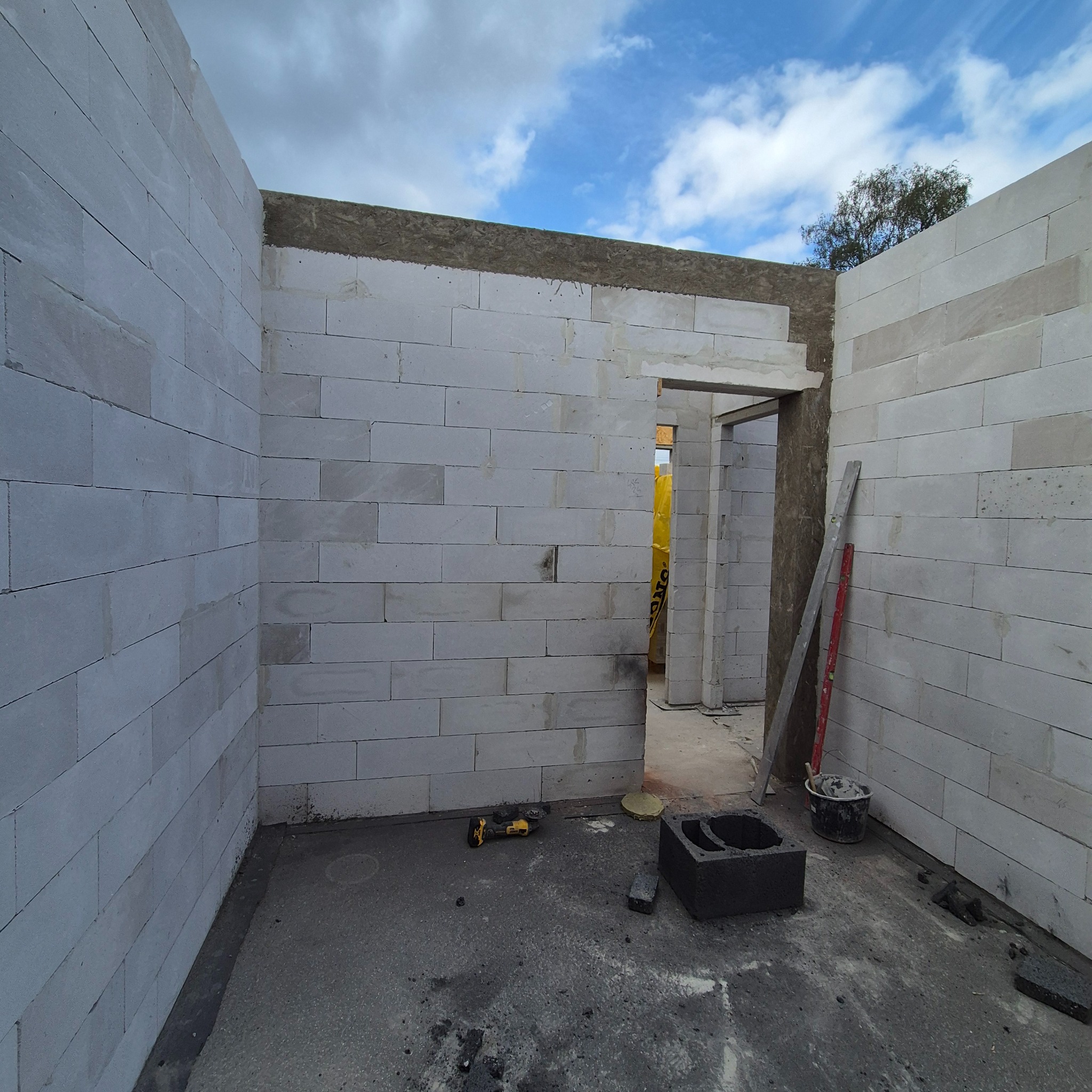 Wnętrze w trakcie budowy: ściany z białych bloczków, widoczne narzędzia (wiertarka), poziomica i wiadro. W tle błękitne niebo. Etap murowania ścian.