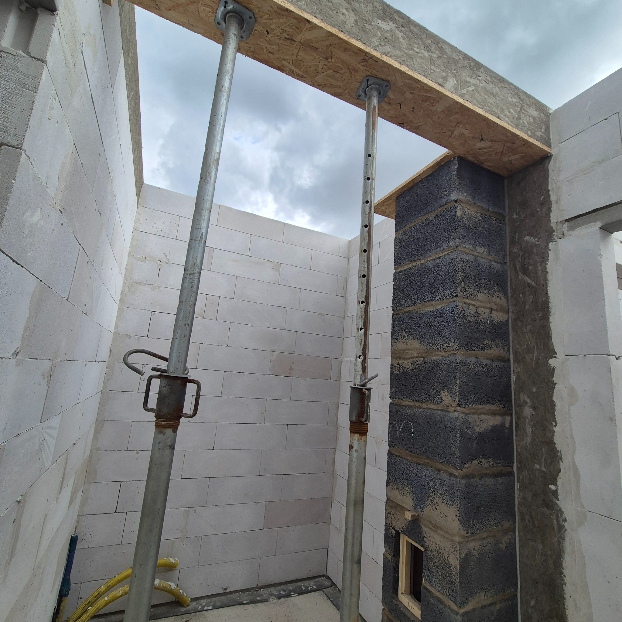 Wnętrze surowego domu z białych bloczków, wspartego metalowymi stemplami i drewnianą belką. Częściowo wymurowany komin z ciemnych bloczków. Widoczne przewody.