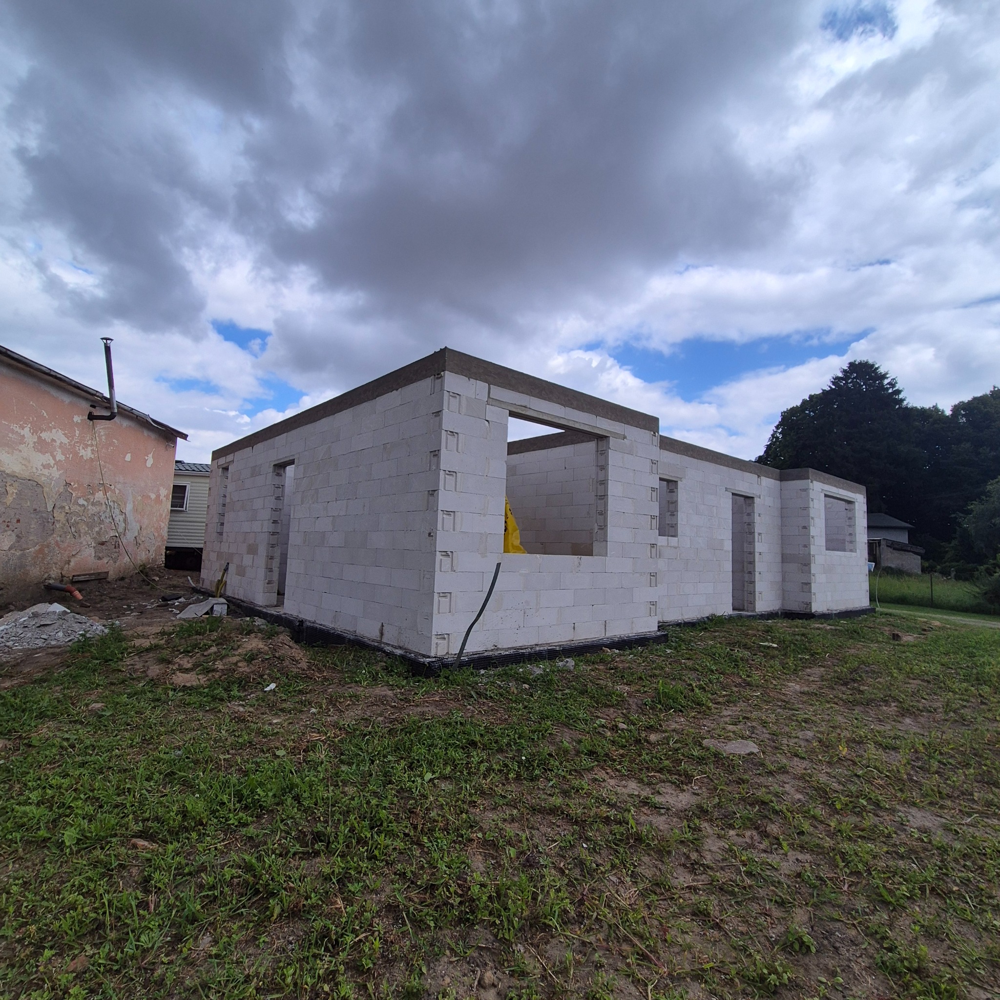 Niski dom z białych bloczków, w trakcie budowy. Widoczne otwory okienne i drzwiowe, szare niebo w tle. Etap murowania ścian.