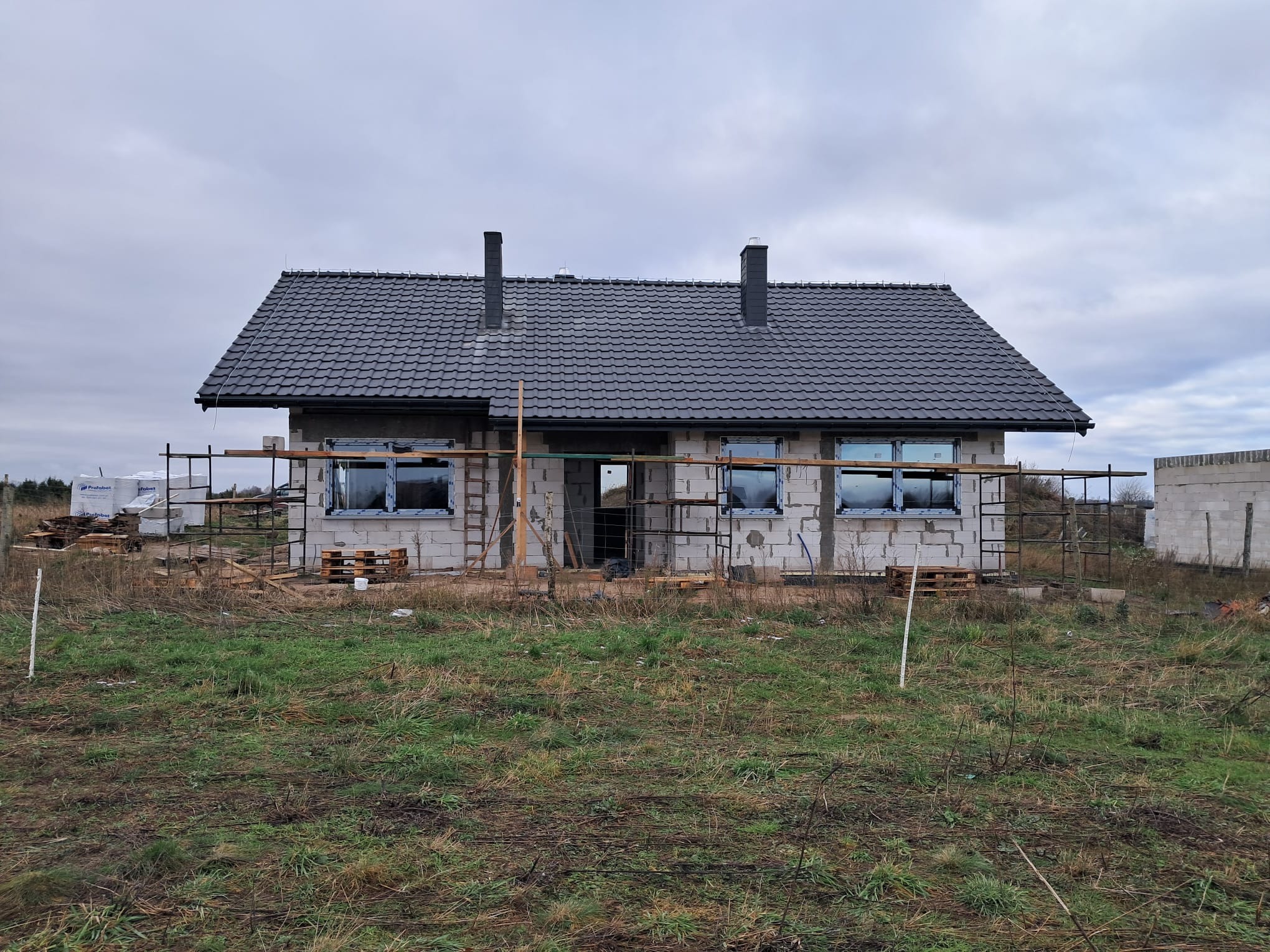 Niedokończony dom z szarej cegły z ciemnym dachem, otoczony rusztowaniami i paletami, na tle pochmurnego nieba i zielonej trawy.