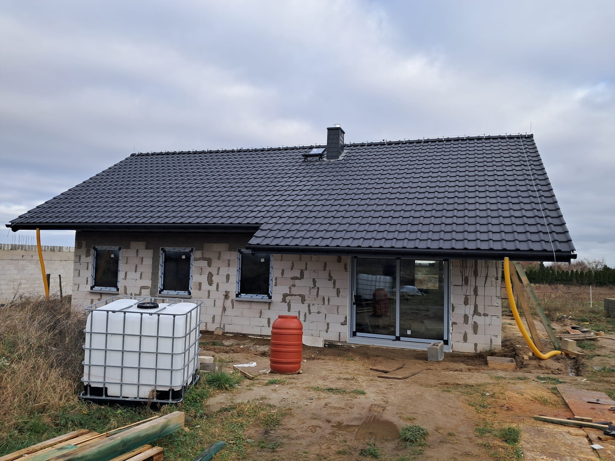 Niedokończony dom z białego pustaka z ciemną dachówką. Widoczne okna, drzwi przesuwne, zbiorniki na wodę i elementy instalacji. Etap budowy w pochmurny dzień.