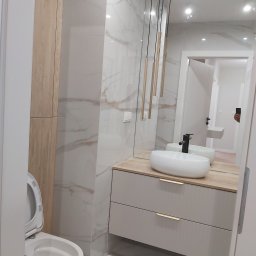 Zakład Ogólnobudowlany - Nowoczesna łazienka z umywalką nablatową na szafce z szufladami, wiszącymi lampami tubami, marmurowymi płytkami na ścianach i wiszącą toaletą z otwartą klapą.