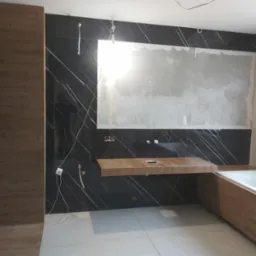 Łazienka w trakcie remontu: czarne płytki z białymi żyłami, drewnopodobna zabudowa wanny i wisząca szafka umywalkowa, ściana częściowo przygotowana pod gładzie.