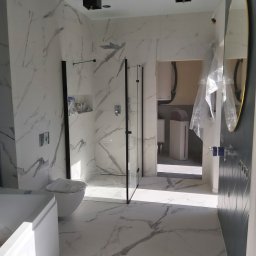 Zakład Ogólnobudowlany - Nowoczesna łazienka z marmurowymi płytkami, kabiną prysznicową walk-in z czarnymi ramami, owalnym lustrem w złotej ramie i czarnymi lampami sufitowymi, widok na pomieszczenie gospodarcze z białą...