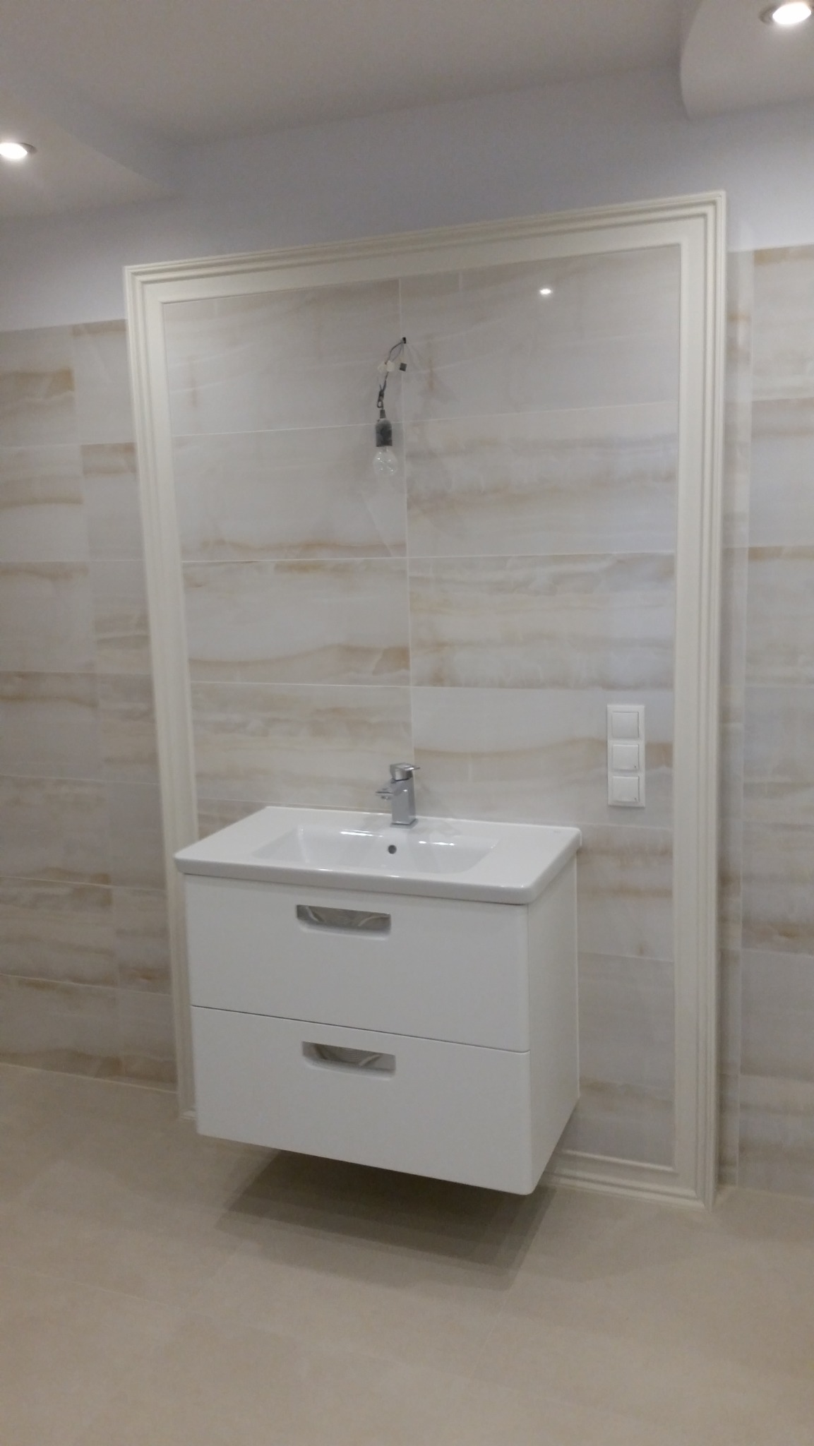 Beżowa łazienka po remoncie: umywalka z szafką w zabudowie, płytki imitujące marmur z dekoracyjną ramą, widoczne przełączniki światła i zwisająca żarówka.