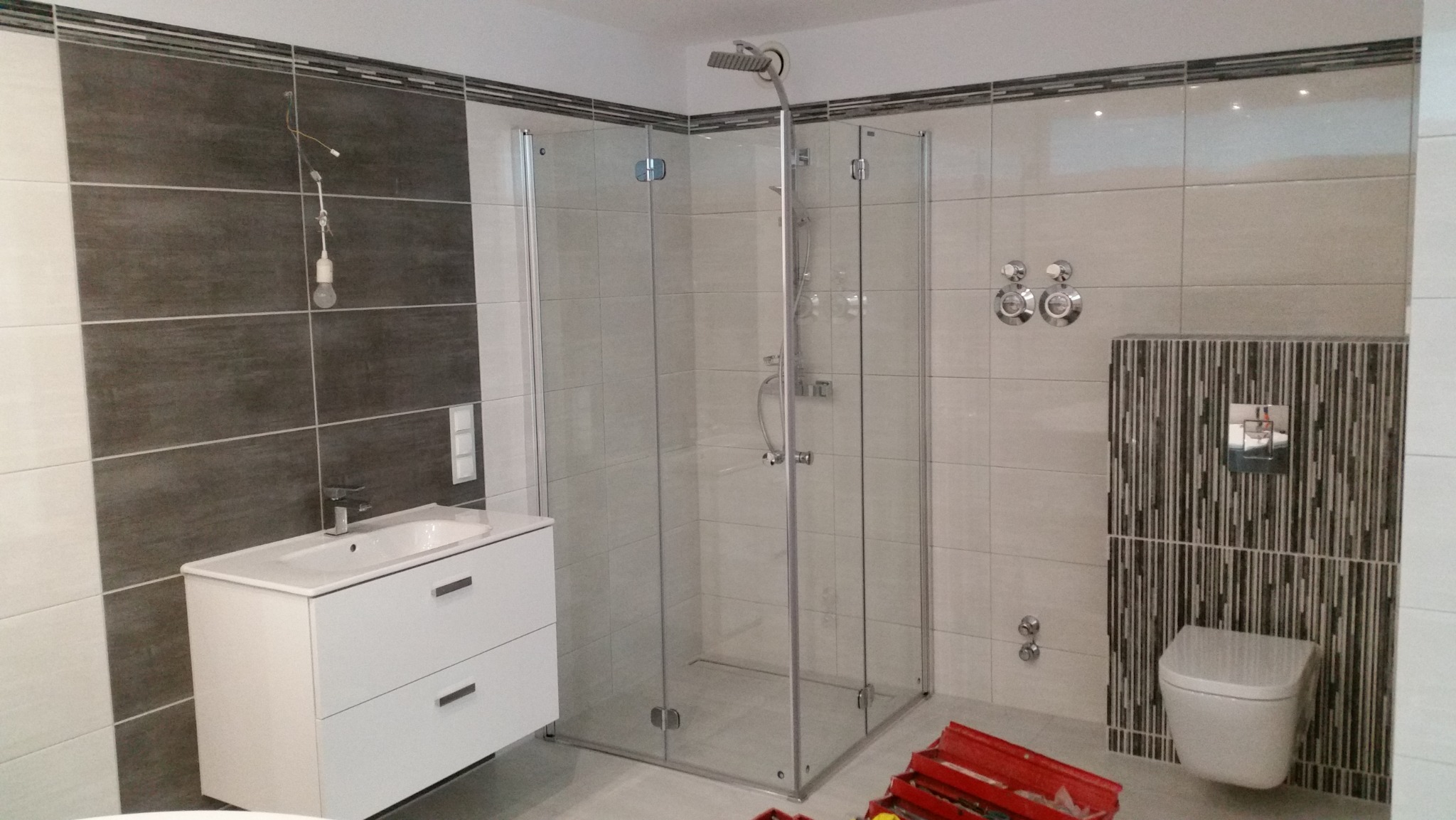 Nowoczesna kabina prysznicowa z białymi i szarymi płytkami w łazience w Lutyni, widoczna umywalka z szafką i częściowo skrzynka z narzędziami na podłodze.