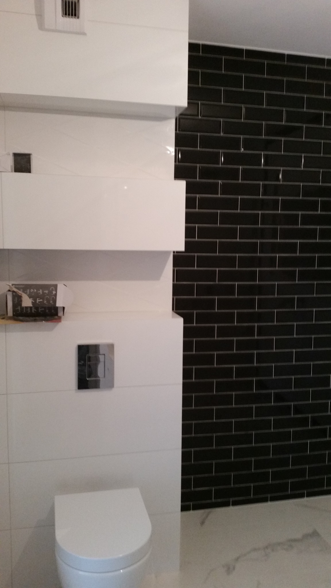 Nowoczesna łazienka z czarną, błyszczącą glazurą w cegiełkę, białymi płytkami i podwieszaną toaletą. Widoczna skrzynka z narzędziami na półce.