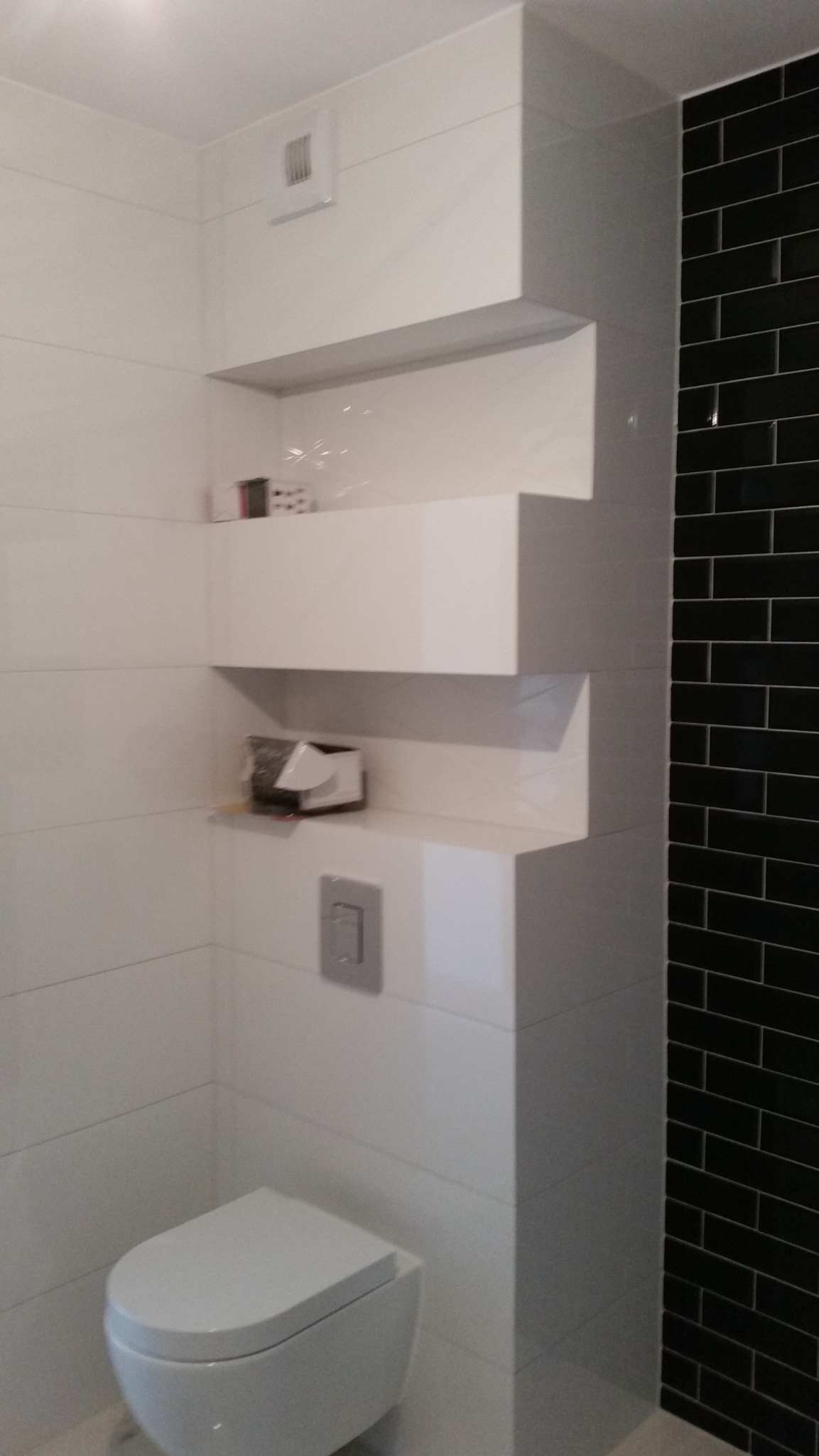 Nowoczesna łazienka z biało-czarnymi płytkami, podwieszaną toaletą i półkami w zabudowie, widoczna kratka wentylacyjna i przycisk spłuczki.