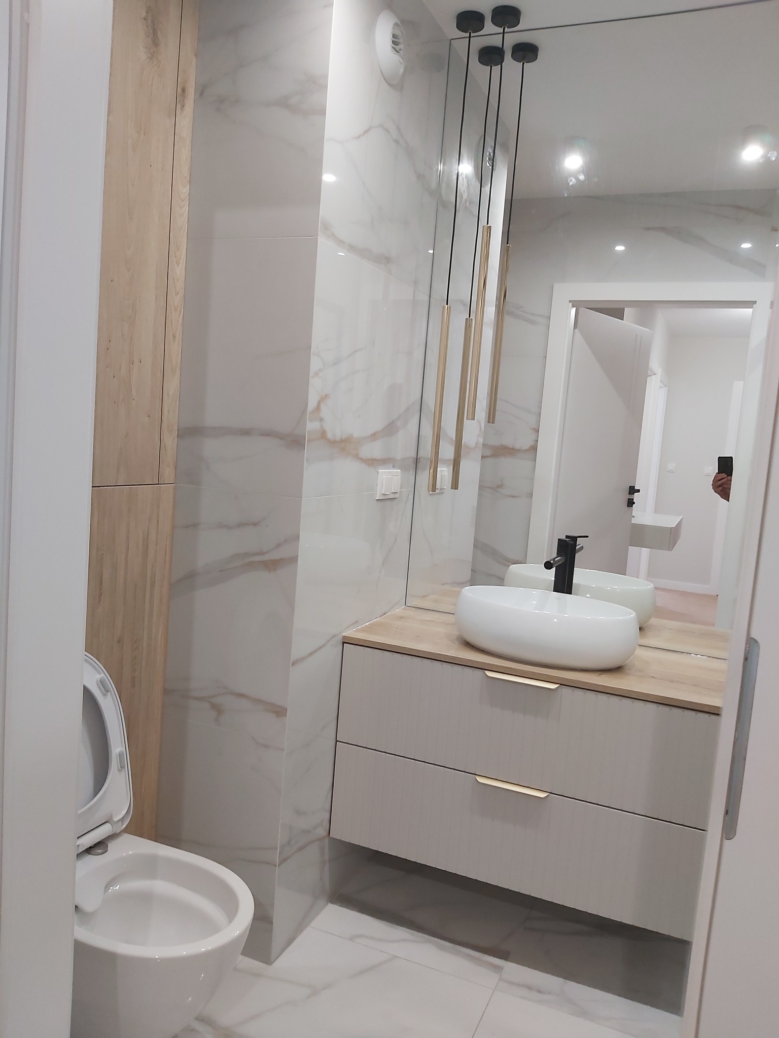Nowoczesna łazienka z umywalką nablatową na szafce z szufladami, wiszącymi lampami tubami, marmurowymi płytkami na ścianach i wiszącą toaletą z otwartą klapą.