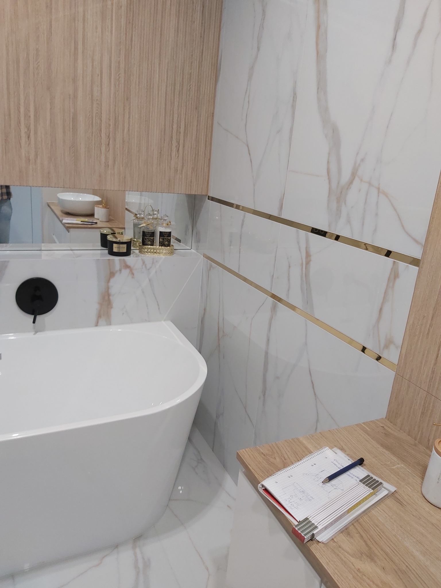 Nowoczesna łazienka z białą, wolnostojącą wanną, armaturą w kolorze czarnym, płytkami imitującymi marmur z złotymi listwami, drewnianymi szafkami i notatnikiem leżącym na blacie.