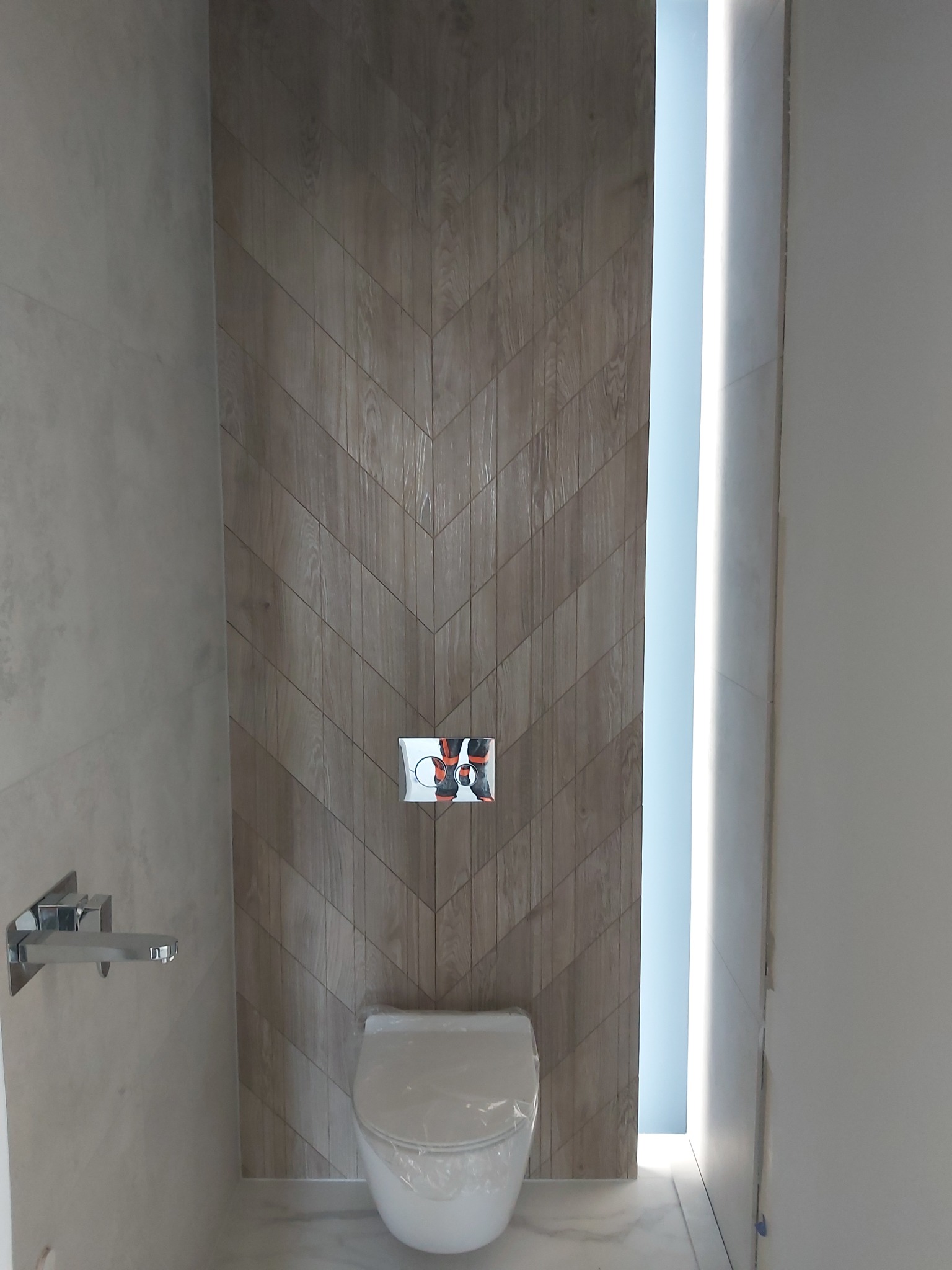 Nowoczesna łazienka z toaletą podwieszaną, drewnopodobne płytki ułożone w jodełkę na ścianie, chromowana bateria umywalkowa i podświetlenie LED w niszy.