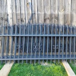 Dominik Gostek - Elementy metalowego ogrodzenia z ozdobnymi detalami, przygotowane do montażu, leżą na trawie przed drewnianym płotem.