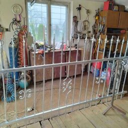 Dominik Gostek - Fragment metalowego ogrodzenia z dekoracyjnymi elementami, wspartego na stojaku w warsztacie z narzędziami i butlami gazowymi w tle.