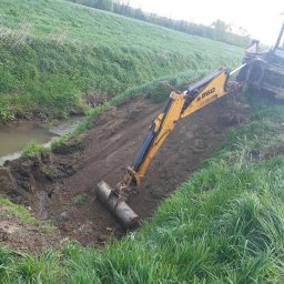 Dominik Gostek - Żółta koparko-ładowarka B60 pogłębia koryto małego cieku wodnego na tle zielonego pola, z widocznym operatorem w jasnej koszulce.