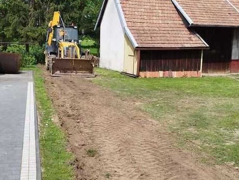 Żółta koparko-ładowarka wyrównuje teren obok budynku gospodarczego z czerwoną dachówką, widoczne ślady opon na świeżo wyrównanej ziemi.