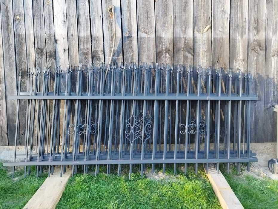 Elementy metalowego ogrodzenia z ozdobnymi detalami, przygotowane do montażu, leżą na trawie przed drewnianym płotem.