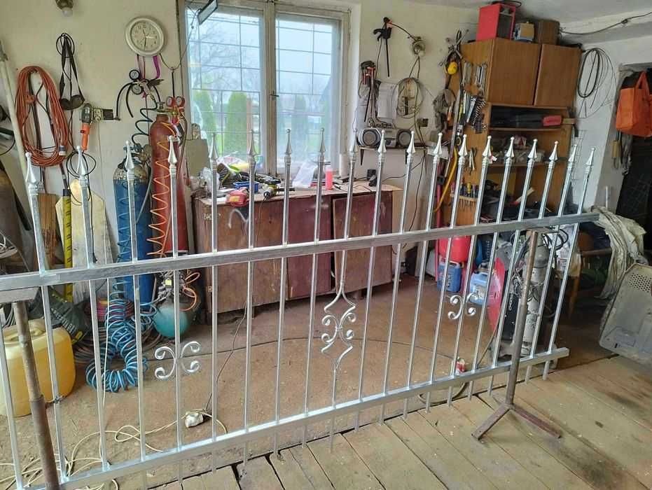 Fragment metalowego ogrodzenia z dekoracyjnymi elementami, wspartego na stojaku w warsztacie z narzędziami i butlami gazowymi w tle.