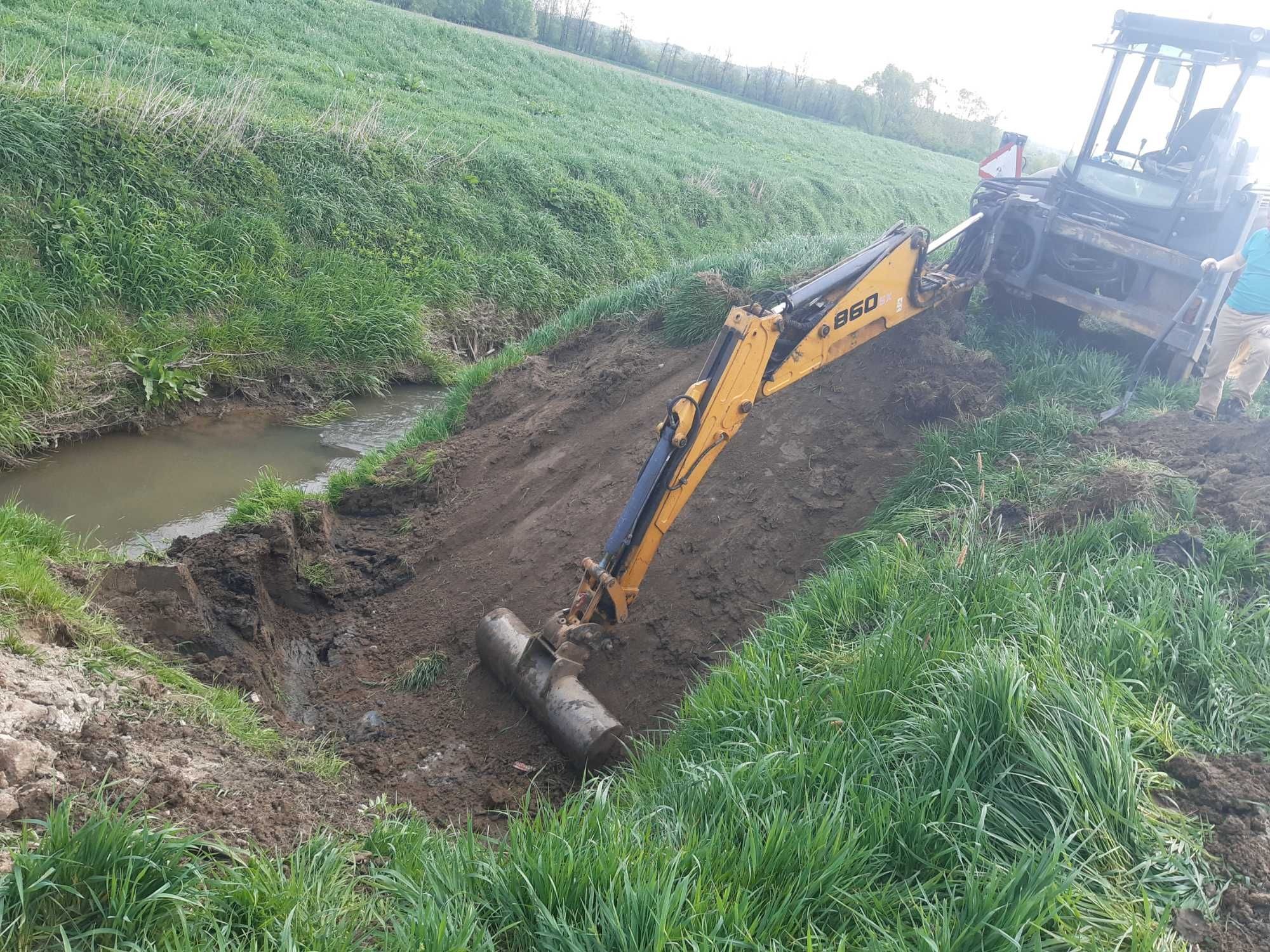 Żółta koparko-ładowarka B60 pogłębia koryto małego cieku wodnego na tle zielonego pola, z widocznym operatorem w jasnej koszulce.