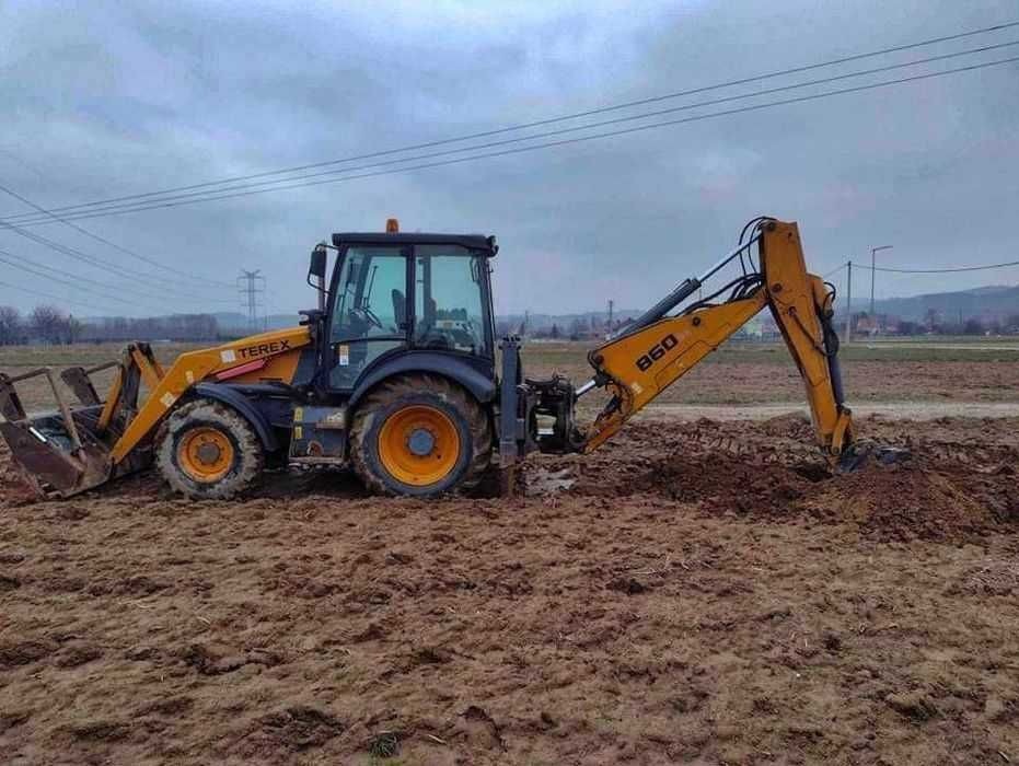 Żółto-czarna koparko-ładowarka Terex 860 w trakcie pracy na błotnistym terenie, z widocznymi śladami opon i świeżo wykopaną ziemią, pochmurne niebo w tle.