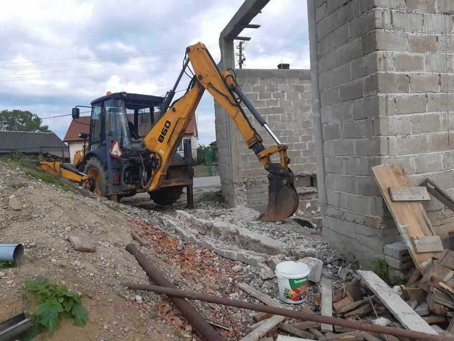 Żółto-czarna koparko-ładowarka B60 częściowo zagrzebana w ziemi, z łyżką skierowaną w stronę ściany z szarego bloczka betonowego, gruz i fragmenty fundamentów w wykopie, w tle budynek mieszkalny.