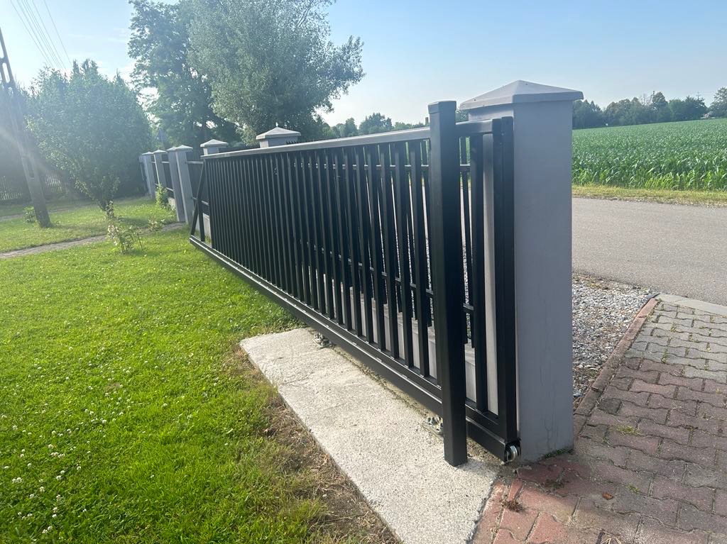 Czarna, przesuwna brama wjazdowa z metalowymi sztachetami, zamontowana na betonowym fundamencie i szarych słupkach, na tle zielonego trawnika i pola uprawnego.