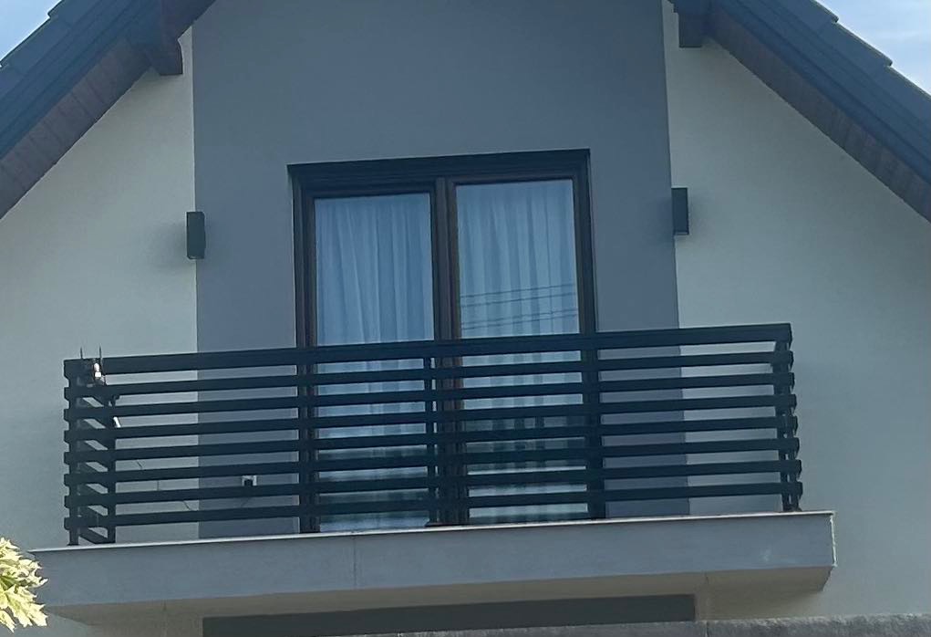 Nowoczesna, czarna, pozioma balustrada balkonowa z metalu, na tle szarej fasady domu jednorodzinnego z oknem balkonowym.