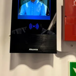 Terminal do pomiaru temperatury i rozpoznawania twarzy Akuvox, zamontowany na ścianie obok czerwonej skrzynki alarmowej. Na ekranie skan twarzy mężczyzny w maseczce.