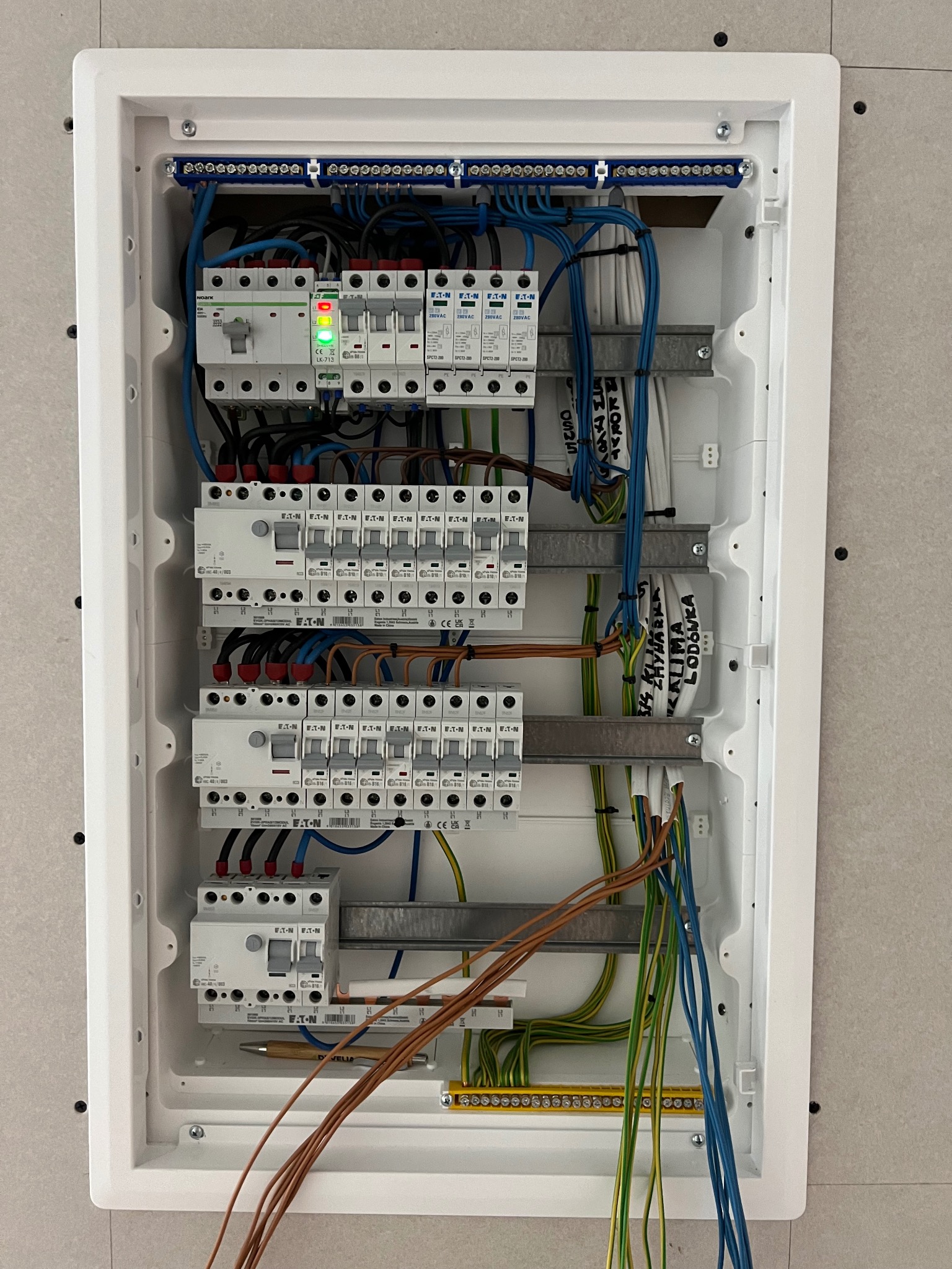 Rozdzielnica elektryczna w białej obudowie, widoczne bezpieczniki, przewody w różnych kolorach (niebieski, zielono-żółty, brązowy), listwy zaciskowe, opisane kable, jeden z wyłączników z zieloną...