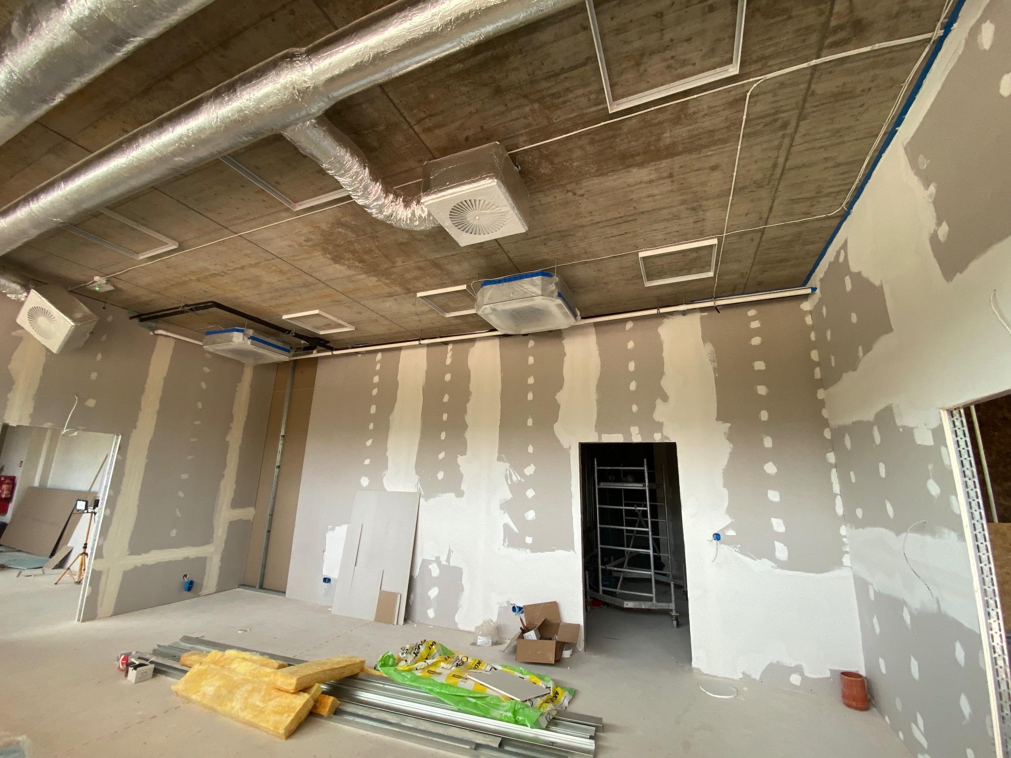 Wnętrze w trakcie wykończenia: surowy betonowy sufit z widocznymi rurami wentylacyjnymi i oprawami oświetleniowymi, ściany z częściowo nałożonym gipsem, materiały izolacyjne i profile metalowe...