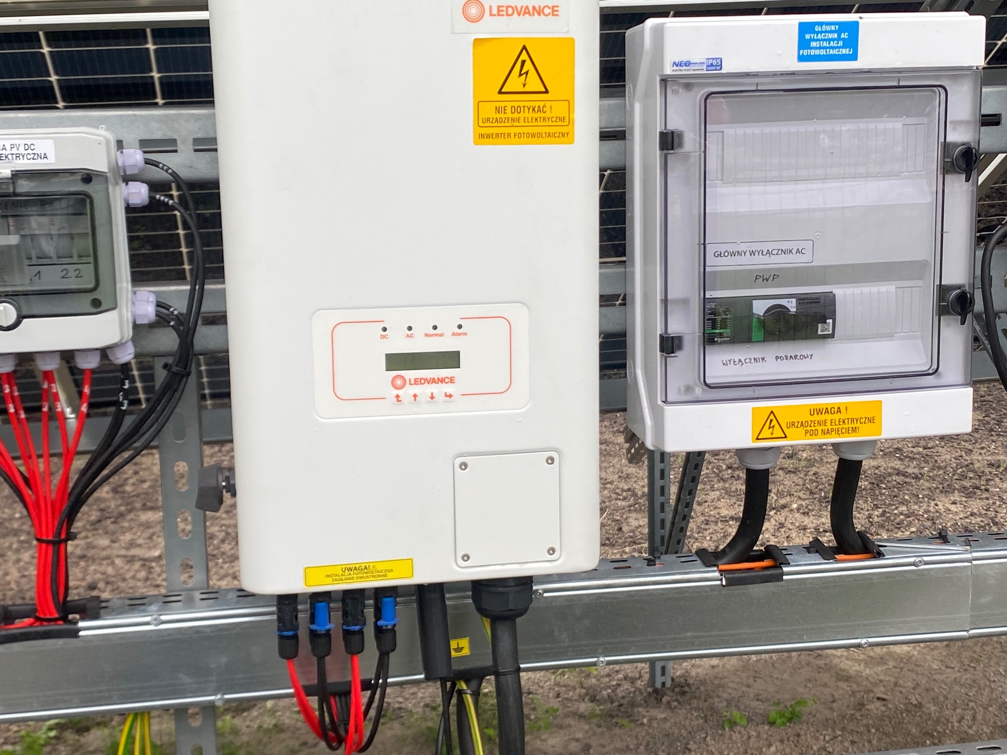 Biała obudowa inwertera fotowoltaicznego Ledvance z żółtą naklejką ostrzegawczą, obok skrzynka z głównym wyłącznikiem AC, kable w kolorach czerwonym, czarnym i żółto-zielonym, mocowane do szarej...