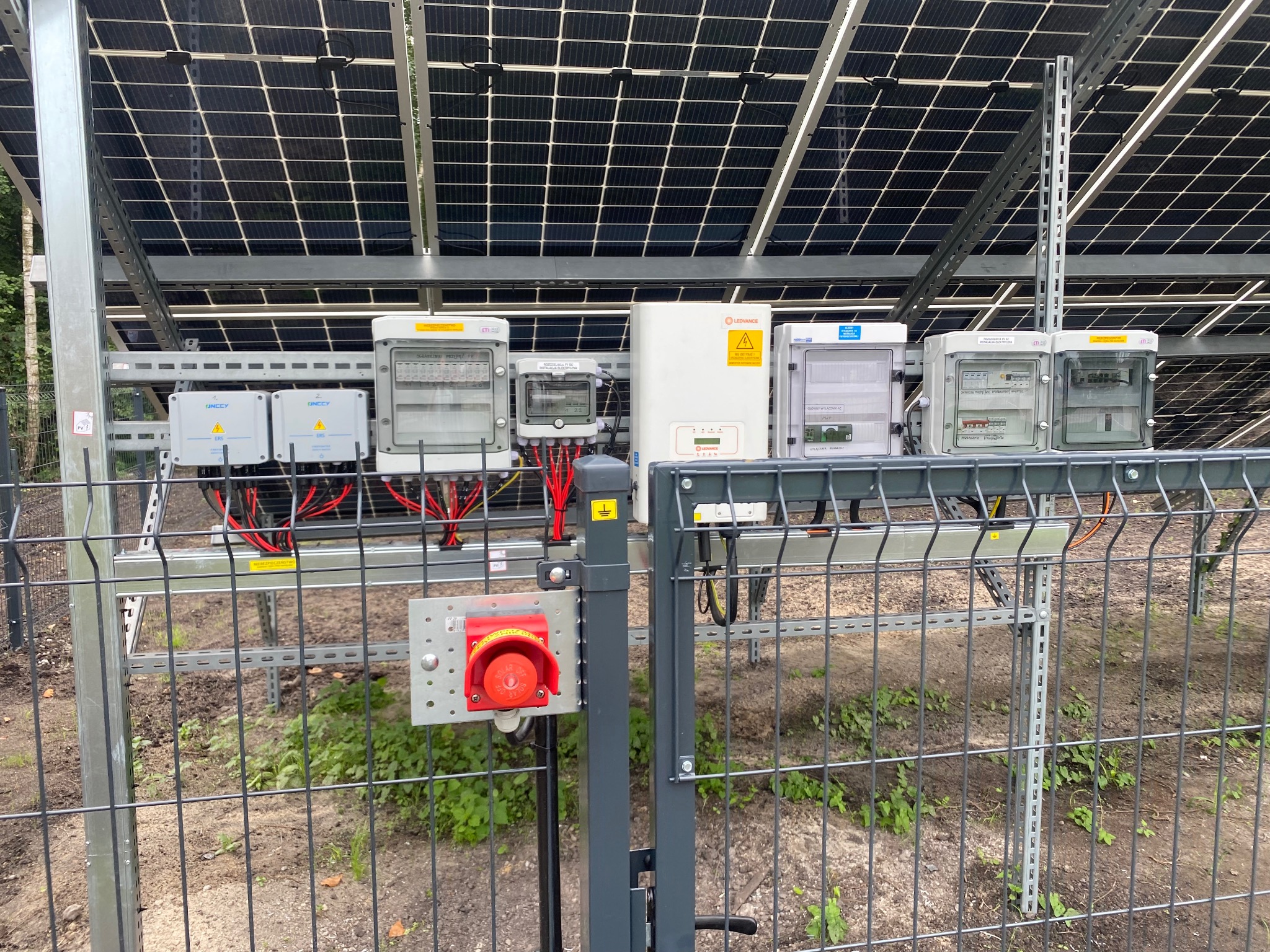 Instalacja paneli fotowoltaicznych z widocznymi zabezpieczeniami, skrzynkami elektrycznymi, inwerterem oraz wyłącznikiem awaryjnym umieszczonym na ogrodzeniu.