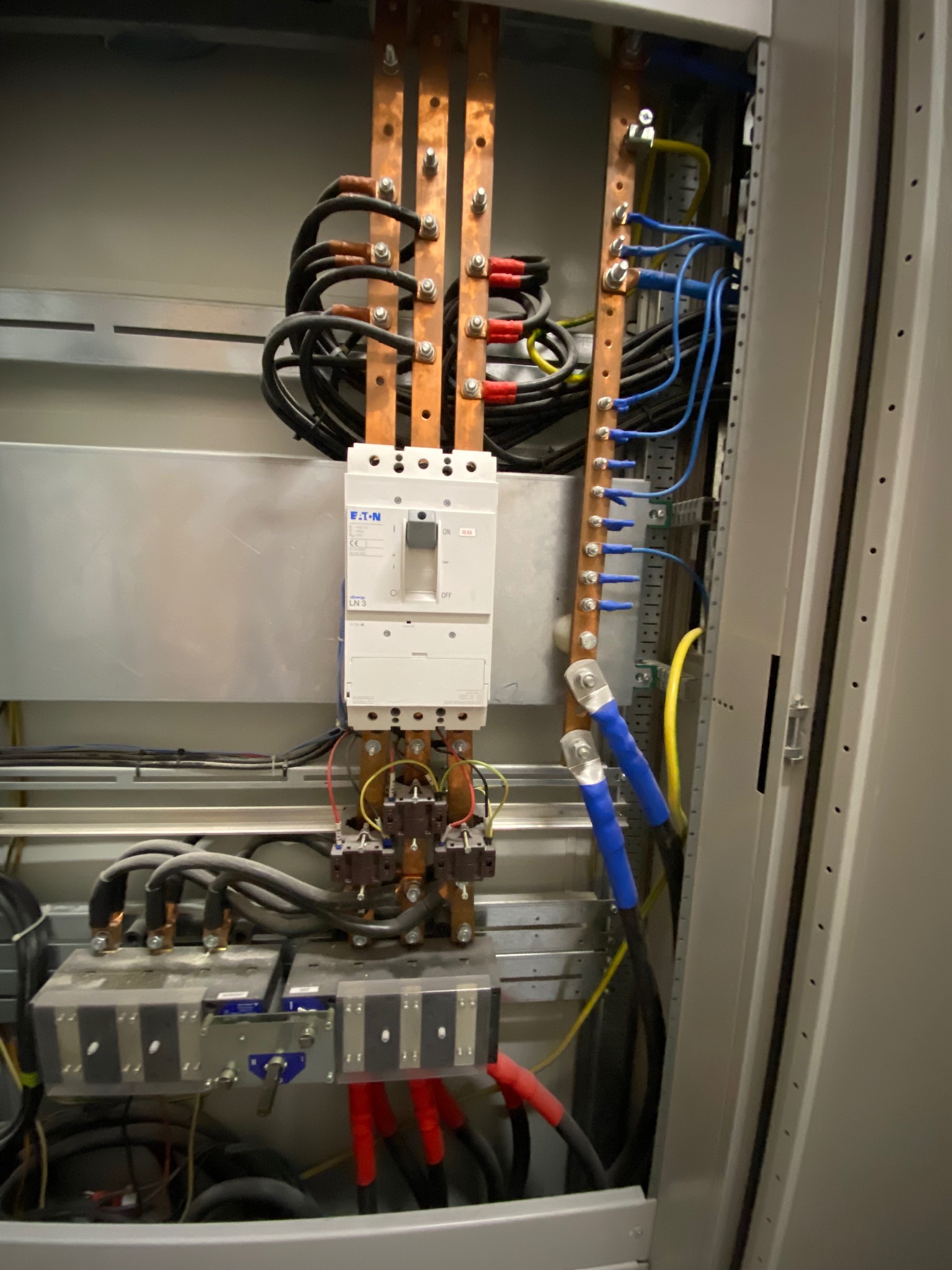 Rozdzielnica elektryczna z szynami miedzianymi, bezpiecznikiem EATON i licznymi przewodami w kolorach czarnym, niebieskim, czerwonym i żółto-zielonym, widoczna wewnątrz metalowej szafy.