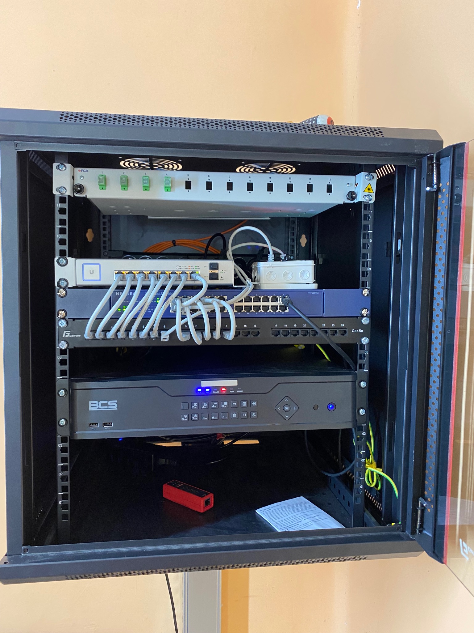 Szafa rack z zainstalowanymi urządzeniami sieciowymi: patch panel, switche, rejestrator BCS, okablowanie strukturalne. Widoczne przewody ethernetowe Cat.5e, tester kabli i instrukcja obsługi.