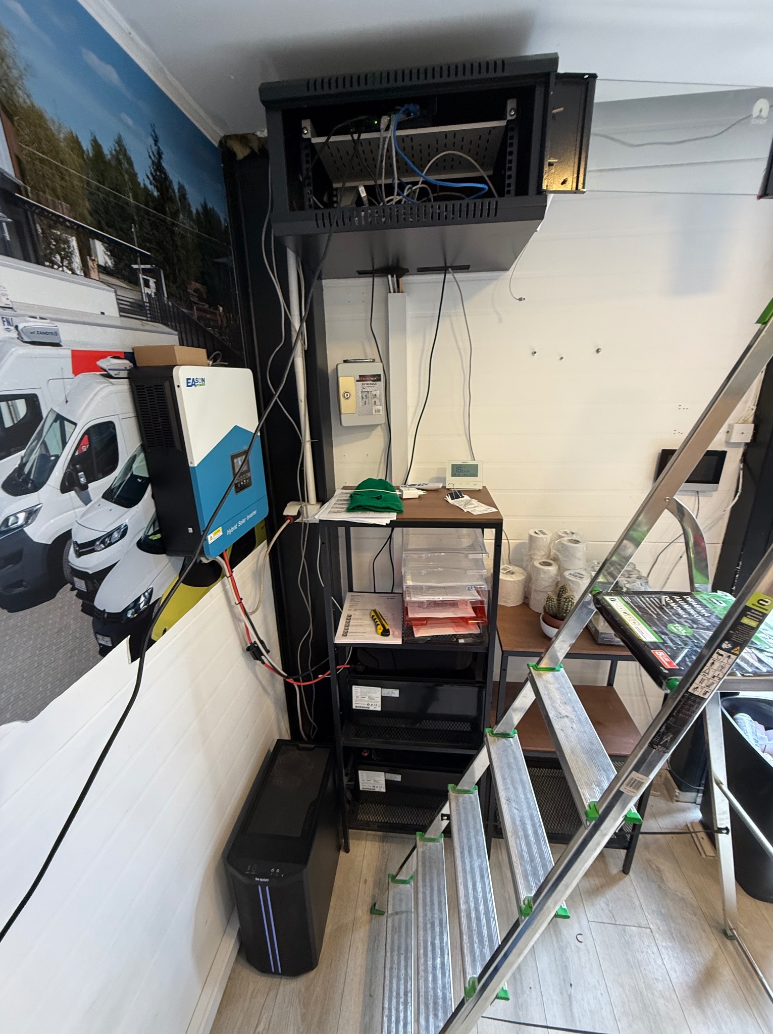 Szafa rack z okablowaniem sieciowym, inwerter solarny EASUN POWER zamontowany na ścianie z fototapetą przedstawiającą samochody dostawcze, obok drabina aluminiowa i UPS be quiet!