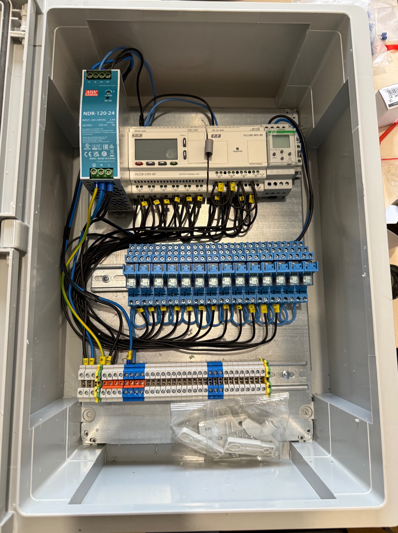 Rozdzielnica elektryczna w szarej obudowie z modułem zasilającym NDR-120-24, programowalnym sterownikiem logicznym FLC18E-8D1-BR oraz modułem przekaźnikowym FLC18-12DI-6R, widoczne okablowanie...