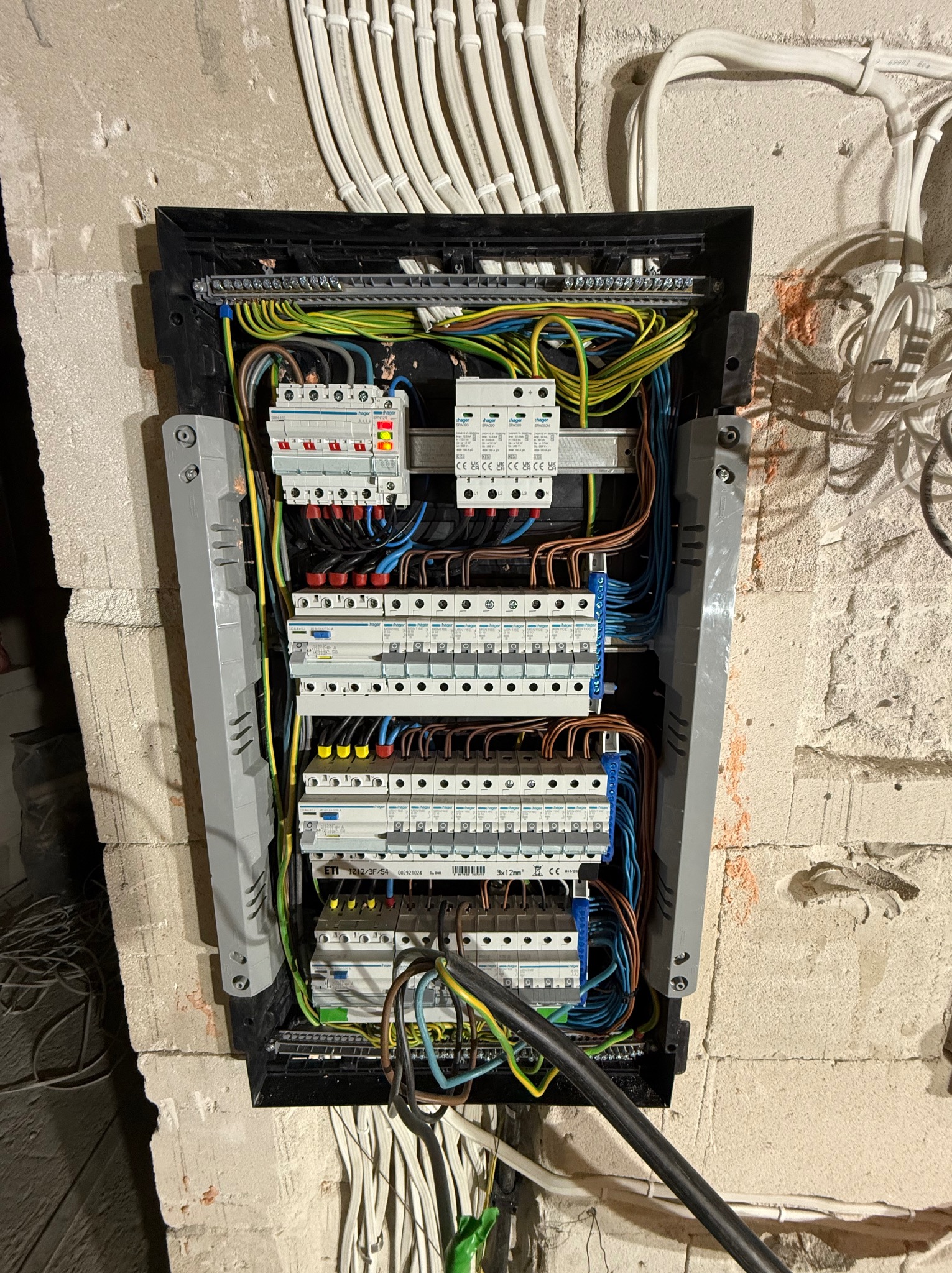 Rozdzielnica elektryczna Hager z licznymi zabezpieczeniami i podłączeniami kablowymi, zamontowana na surowej ścianie z betonu komórkowego. Widoczne przewody w peszlach.